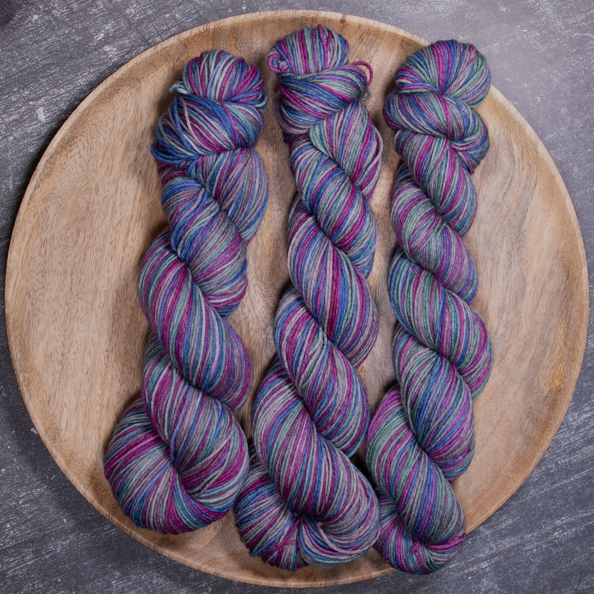 Drei Stränge gesprenkeltes Garn in Blau, Lila und Grün auf einem Holzteller.