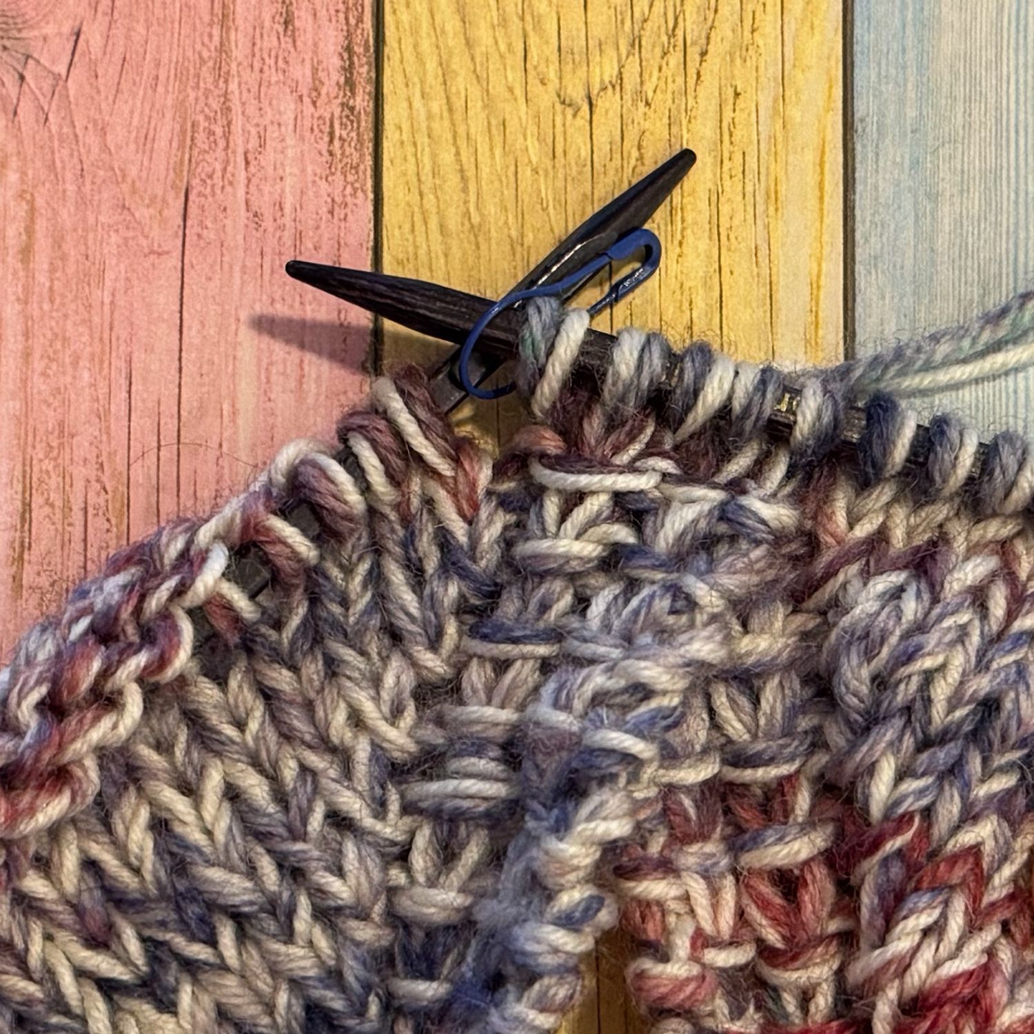 Gestrickter Stoff mit blauem Maschenmarkierer auf farbiger Holzoberfläche.