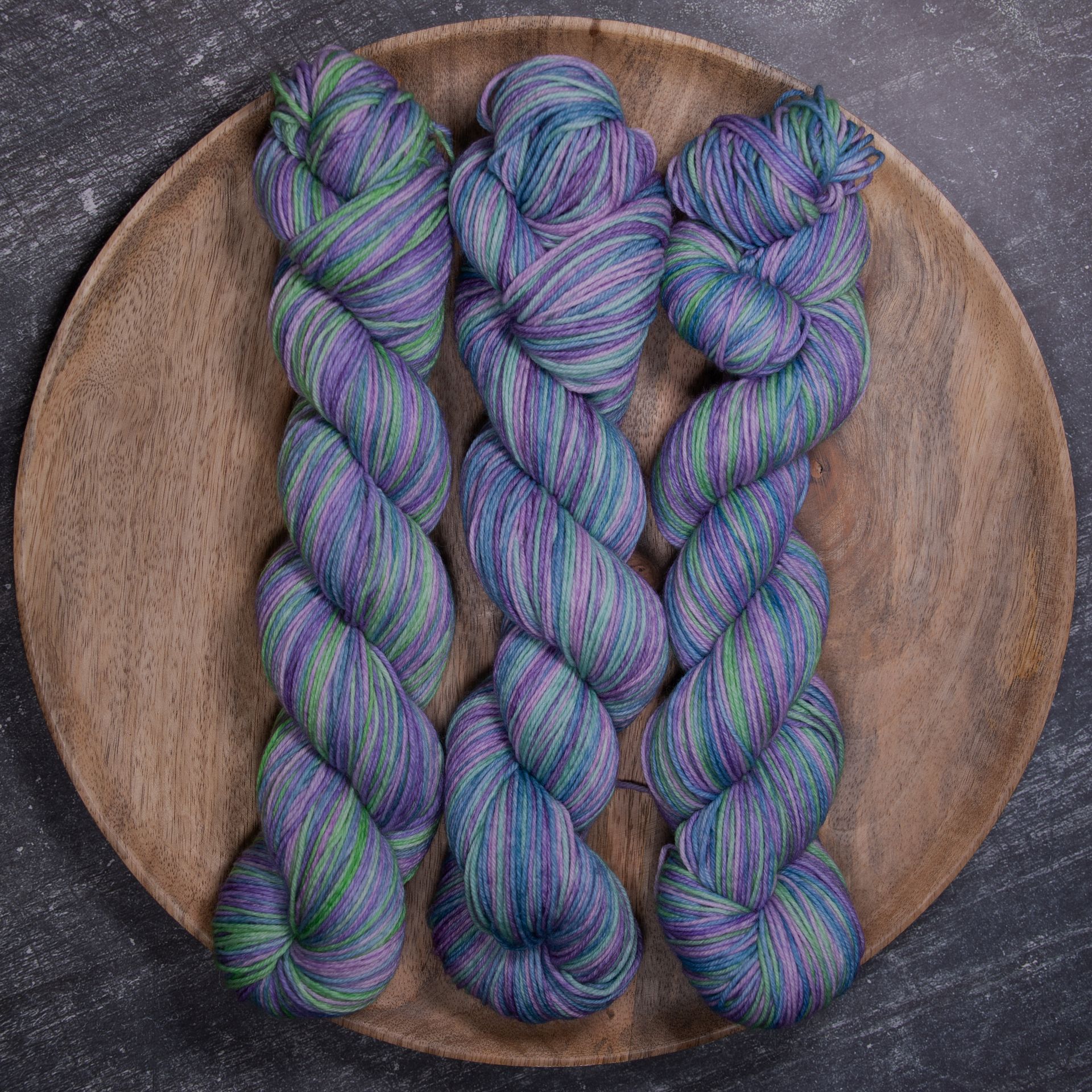 Drei Stränge gesprenkeltes Garn in Blau, Lila und Grün auf einem Holzteller.