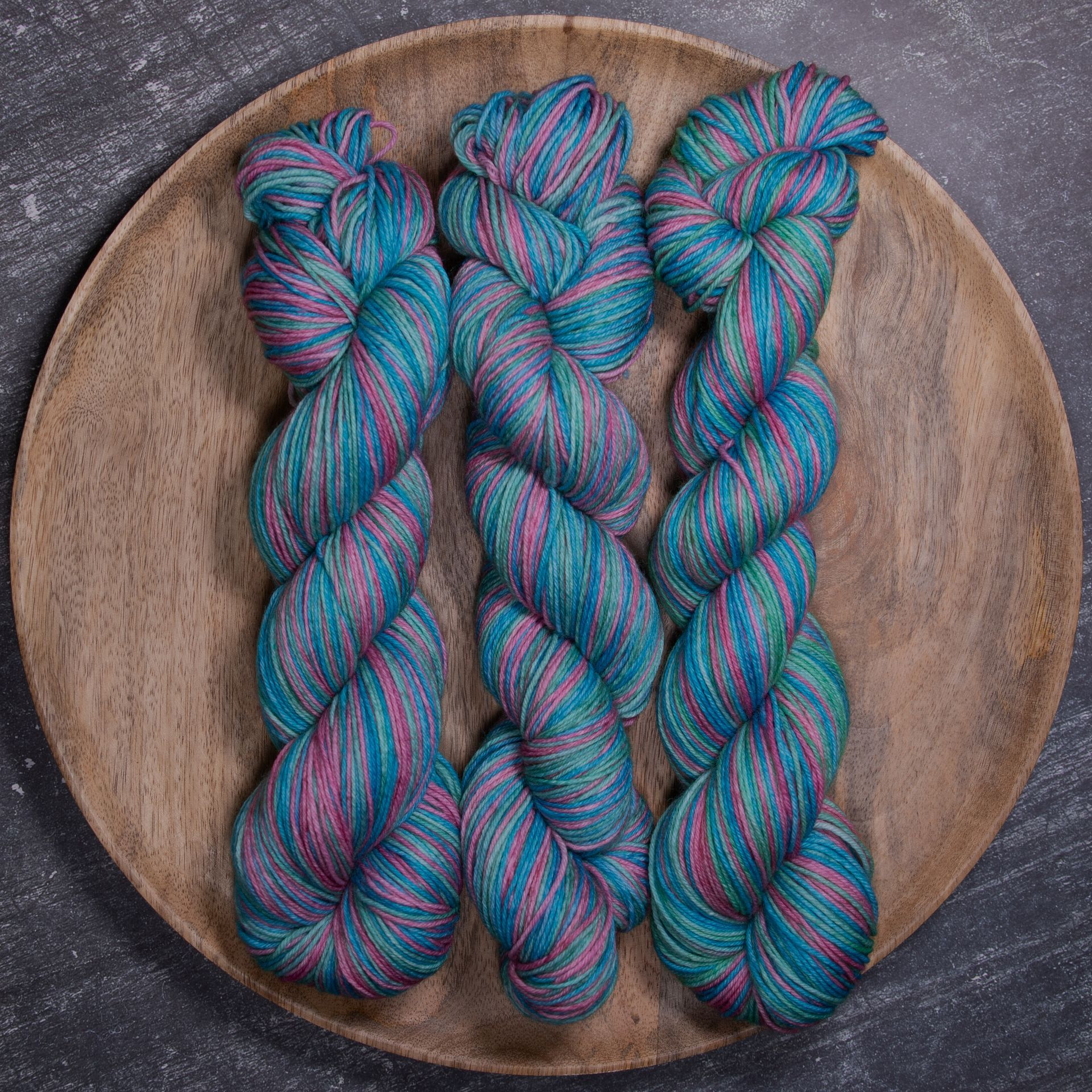 Drei Stränge Garn in den Farben Türkis, Rosa und Grün sind auf einem Holzteller angeordnet.
