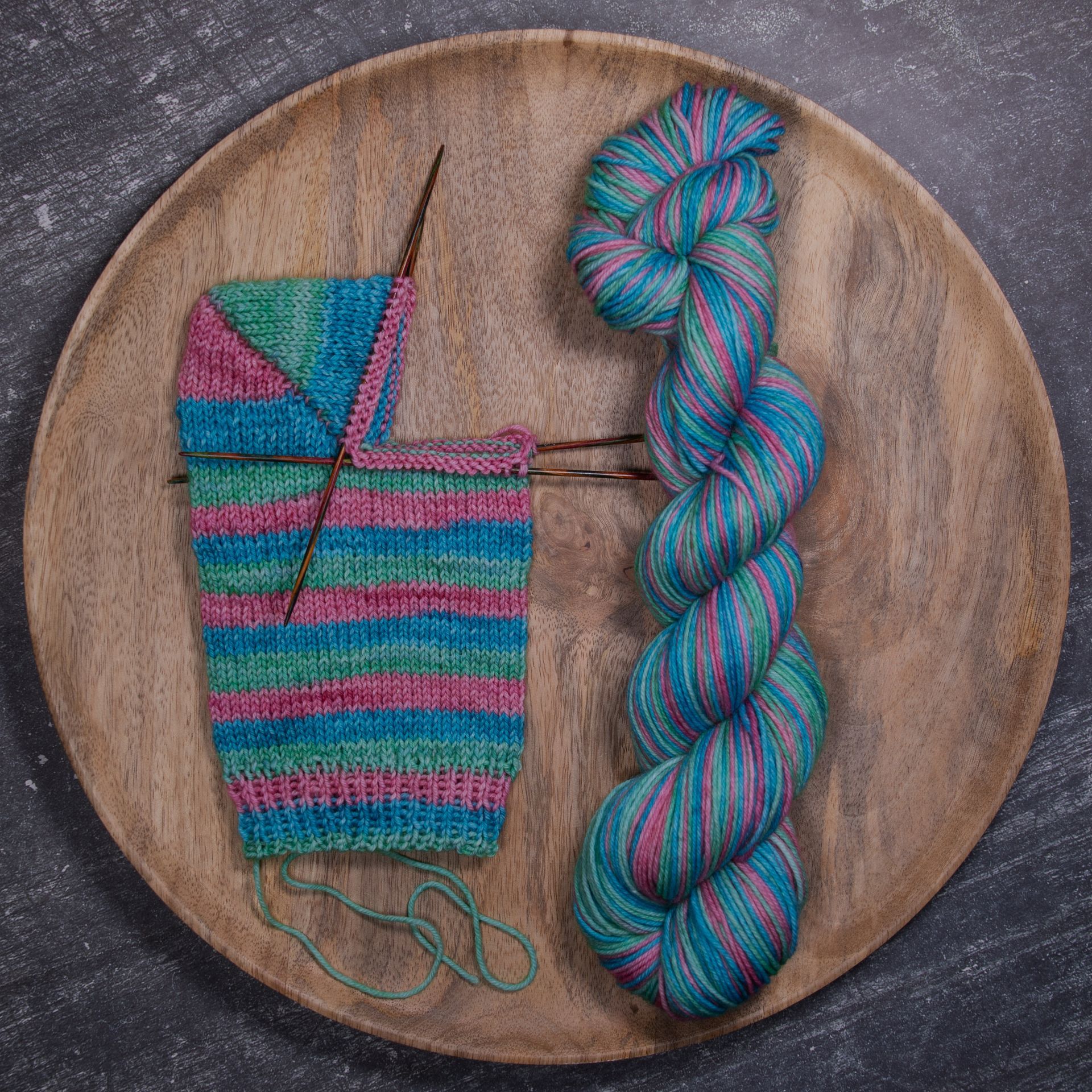 Eine angefangene Stricksocke liegt neben einem Knäuel bunter Wolle auf einem Holztablett.