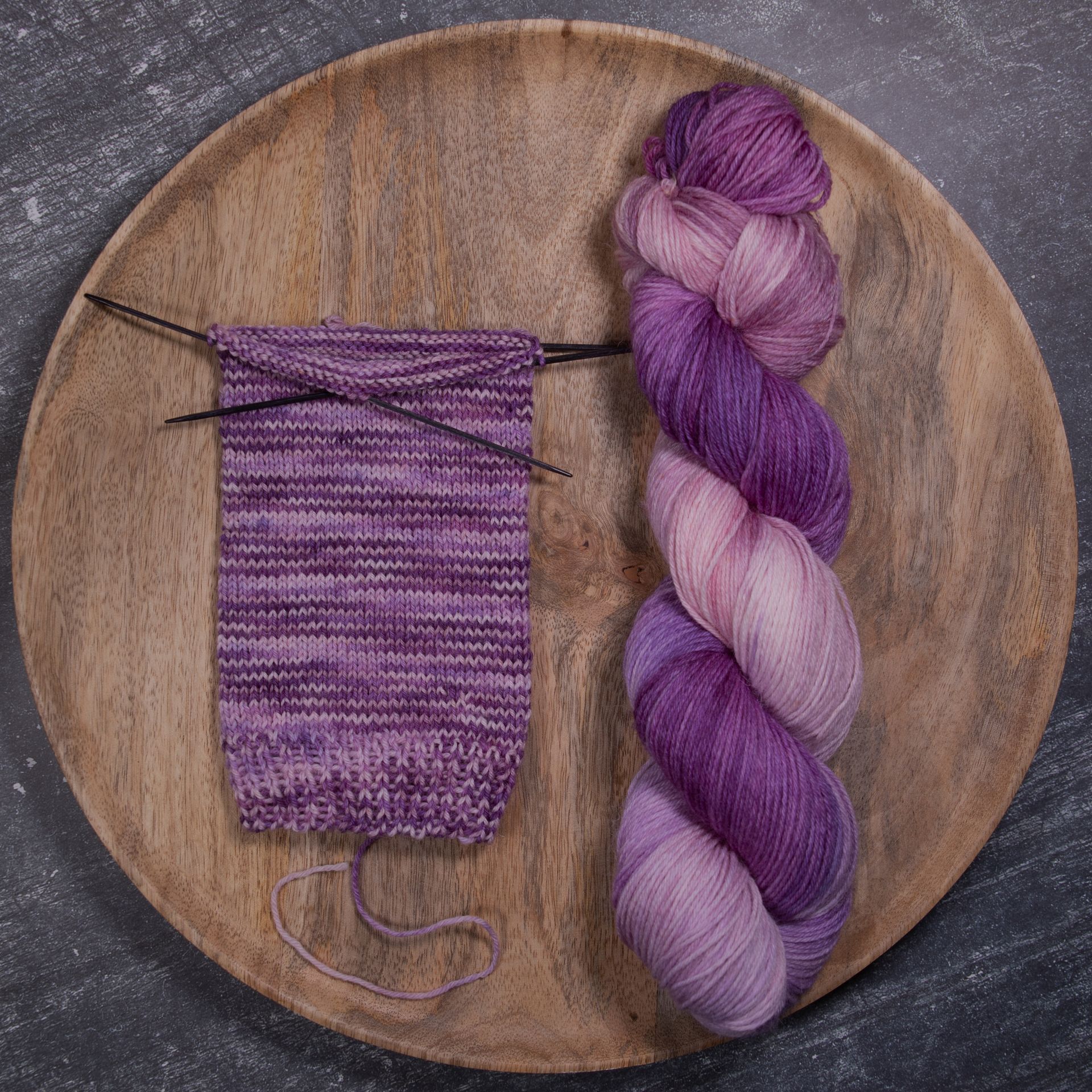 Ein Knäuel lila Garn und eine Strickprobe einer Socke auf einem Holzteller, dazu die Stricknadeln.