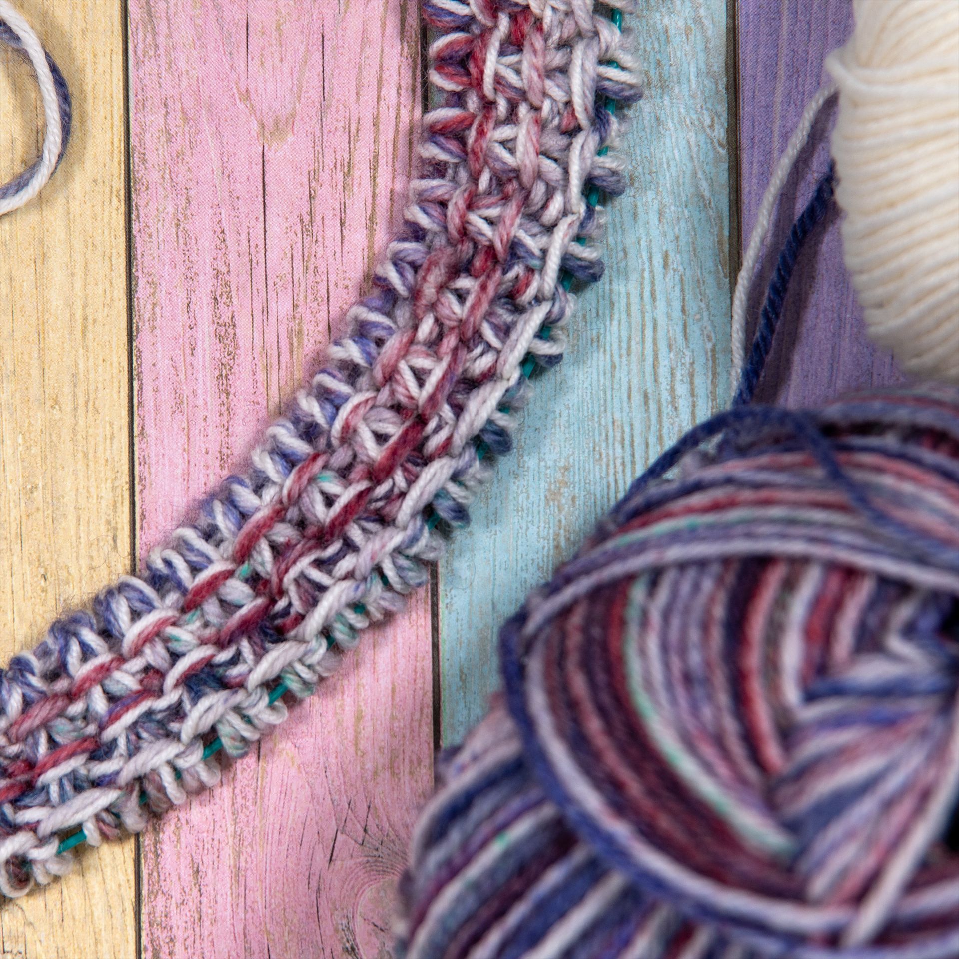 Gestrickter, mehrfarbiger Schal und Wollknäuel auf pastellfarbenem Holzgrund.