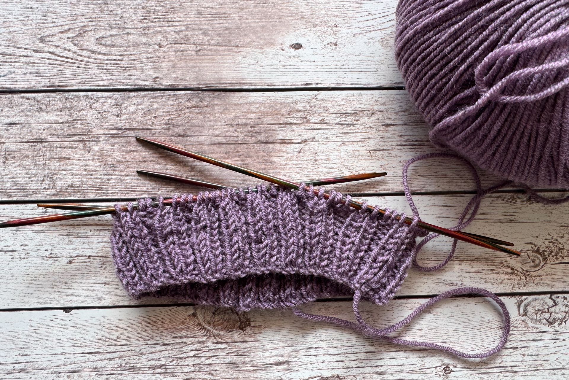 Gestrickter lila Bündchen auf Nadeln, neben einem Wollknäuel, auf einer Holzfläche.