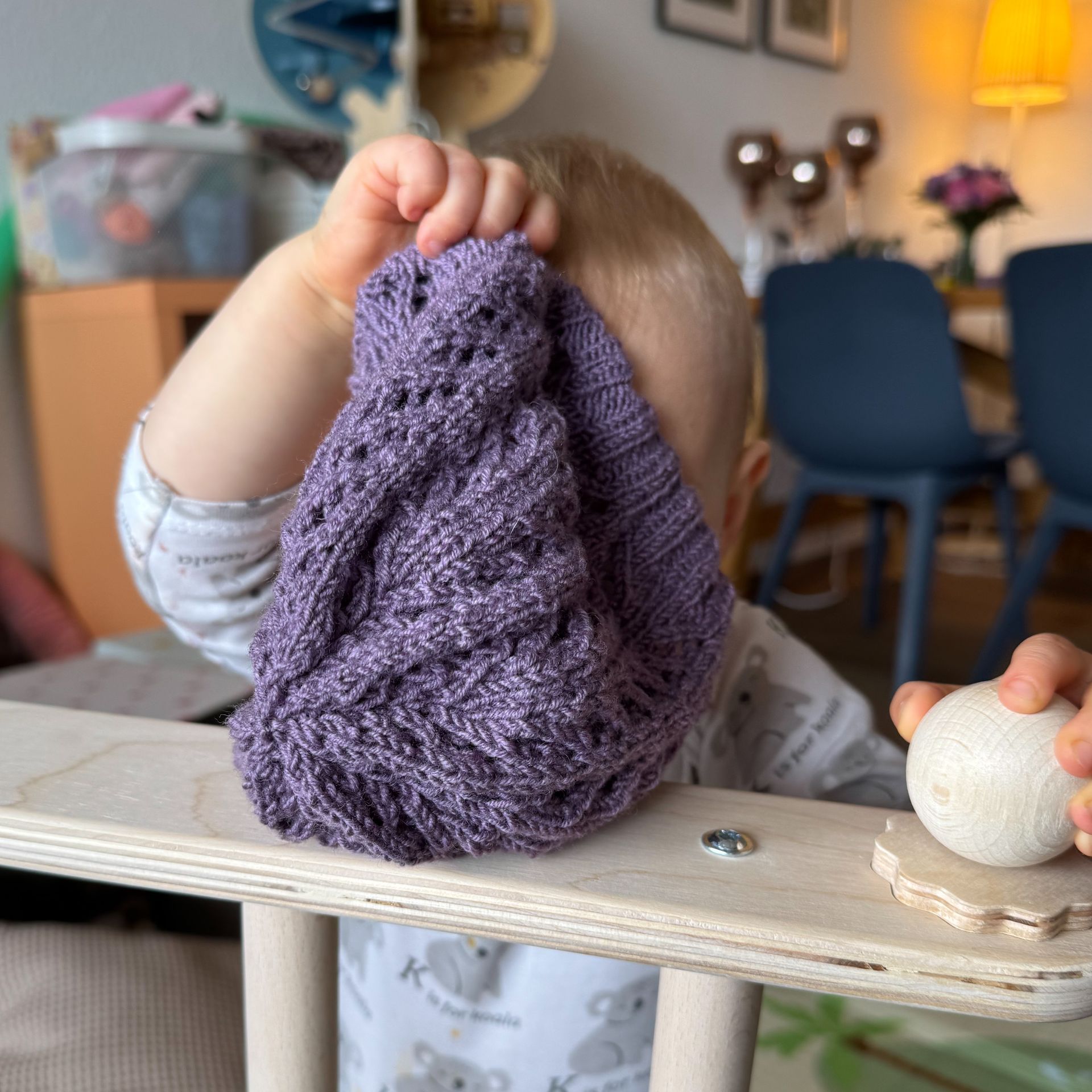 Kind hält eine lila Strickmütze hoch. Sitzt auf einem hölzernen Hochstuhl, verschwommener Hintergrund.