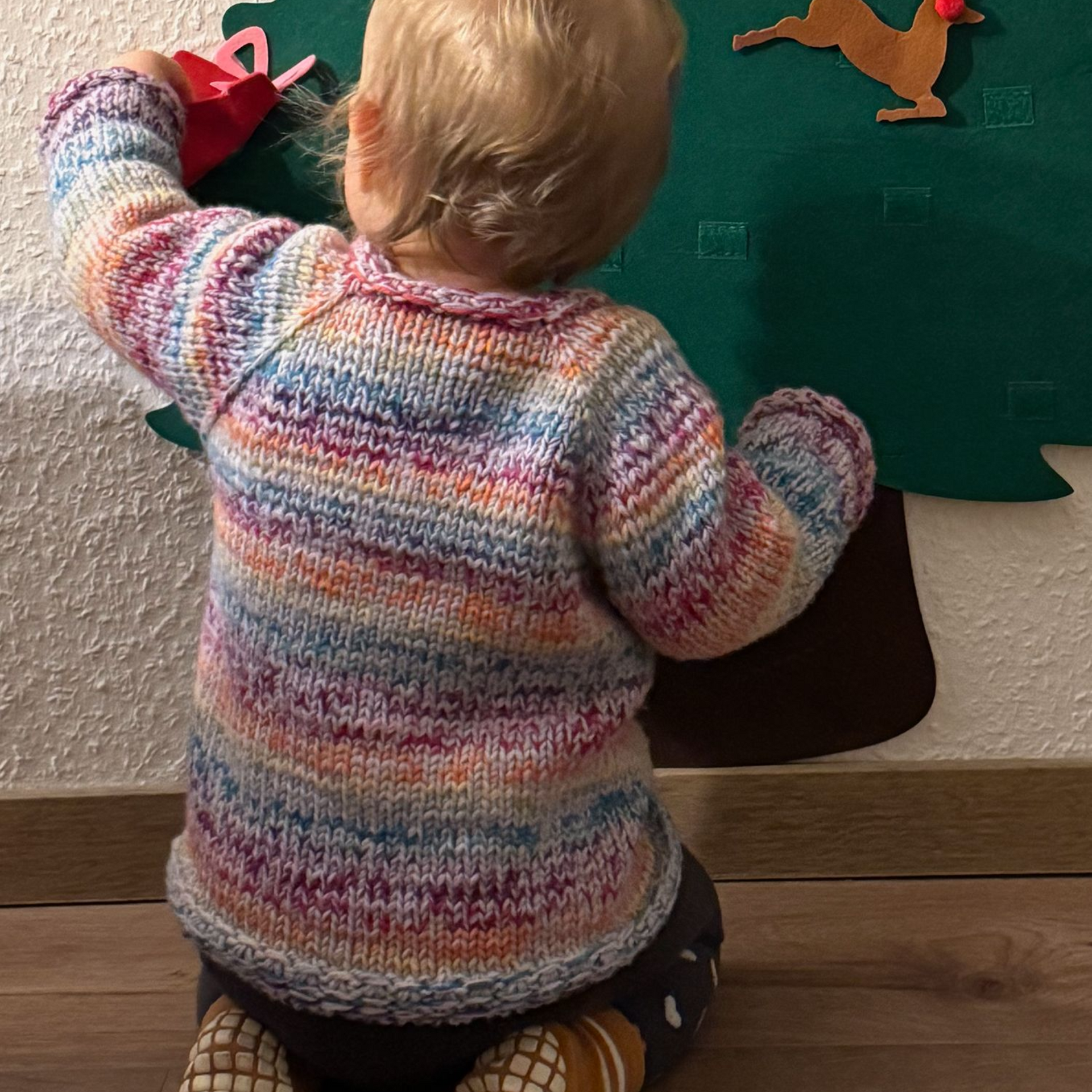 Kind in einem bunten Pullover, kniet neben einem Filzbaum und hält eine rote Gießkanne.