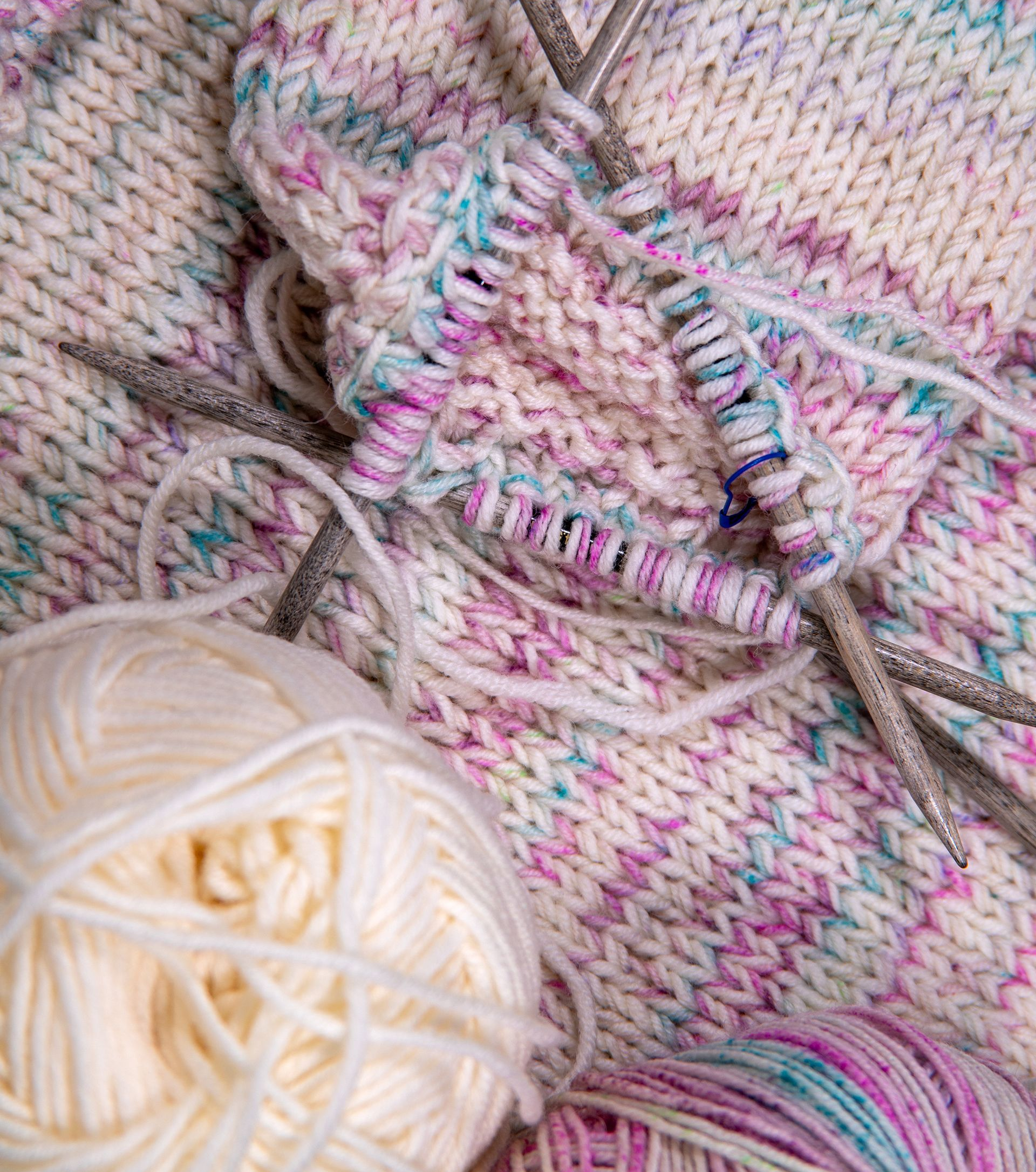 Strickarbeiten mit buntem Garn; Nadeln halten Maschen; cremefarbene und gesprenkelte Pastellgarnknäuel.