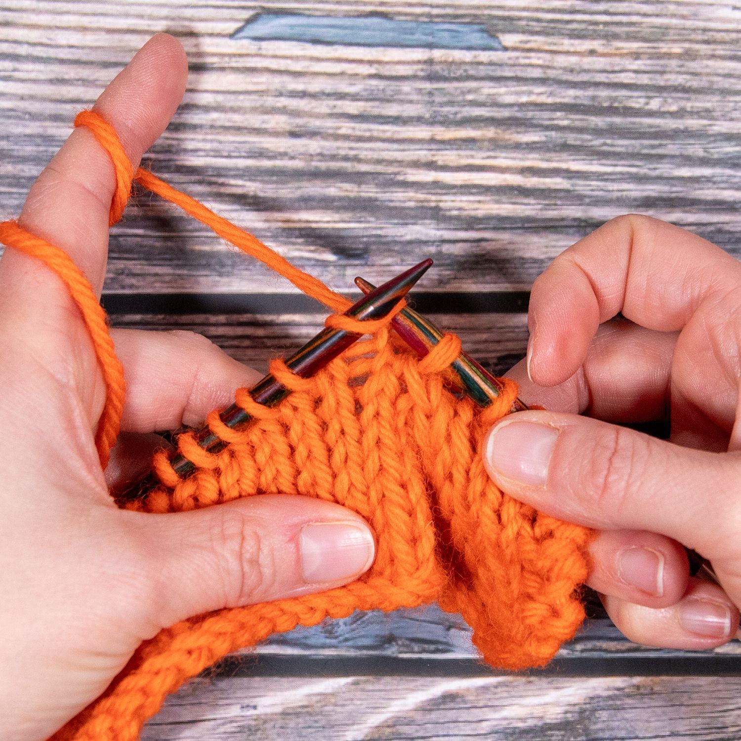 Hände stricken orangefarbenes Garn auf einer Holzfläche. Eine Garnschlaufe liegt auf dem Zeigefinger. Die Stricknadeln sind im Einsatz.