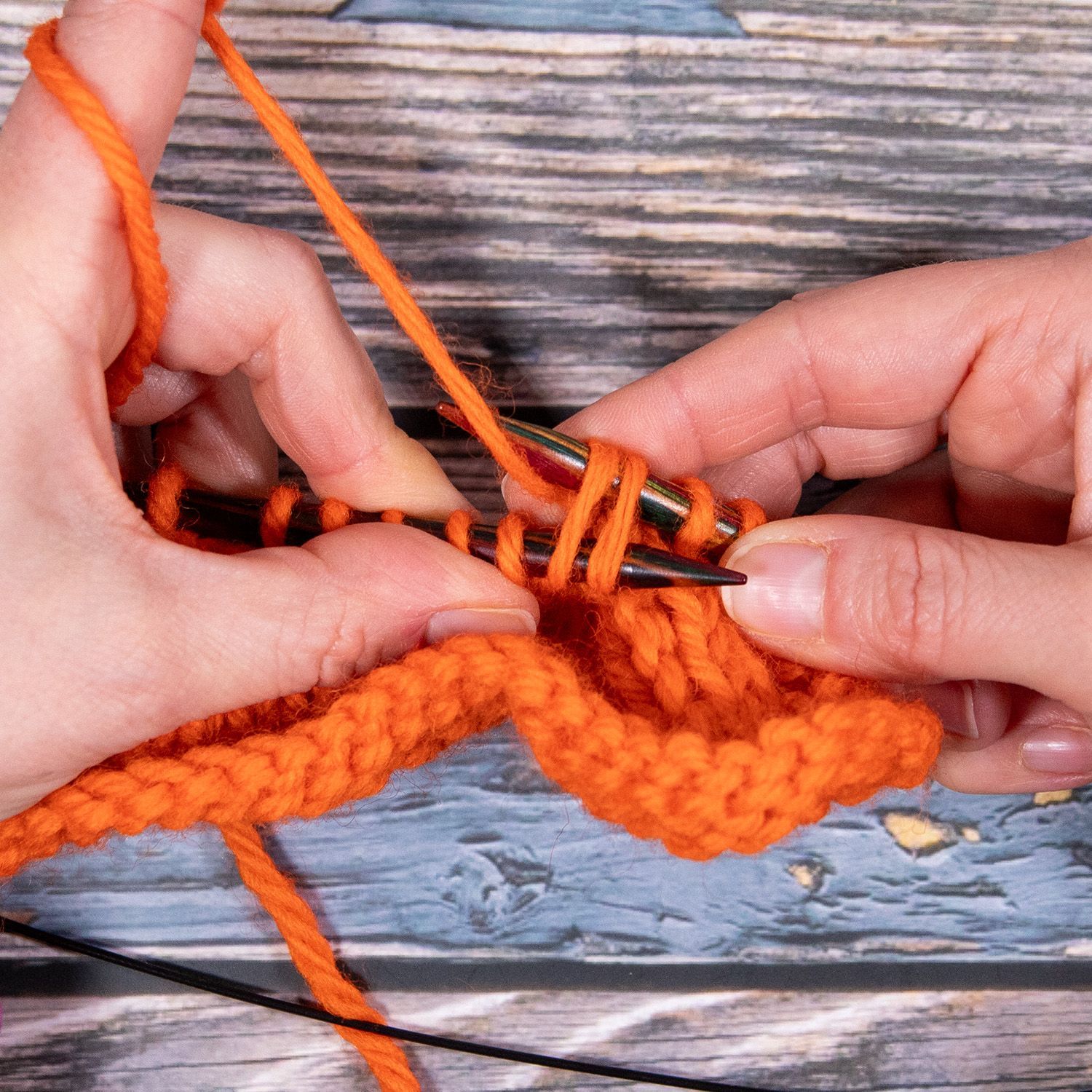 Eine Person strickt mit orangefarbenem Garn auf einer Holzoberfläche. Ihre Hände führen die Stricknadeln, um eine Masche zu erzeugen.