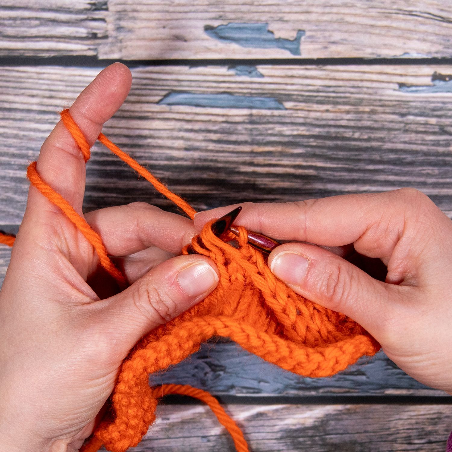 Hände stricken orangefarbenes Garn vor einem Holzhintergrund.