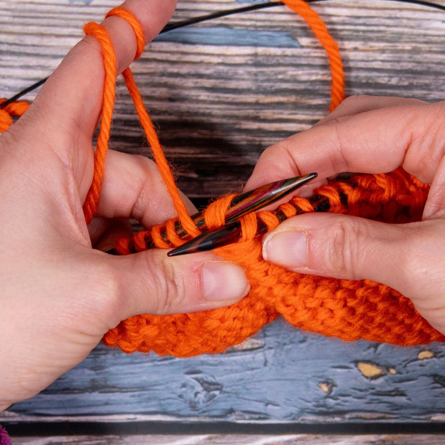 Hände stricken orangefarbenes Garn auf einer Holzoberfläche. Nadeln, Maschen und Garn sind sichtbar.