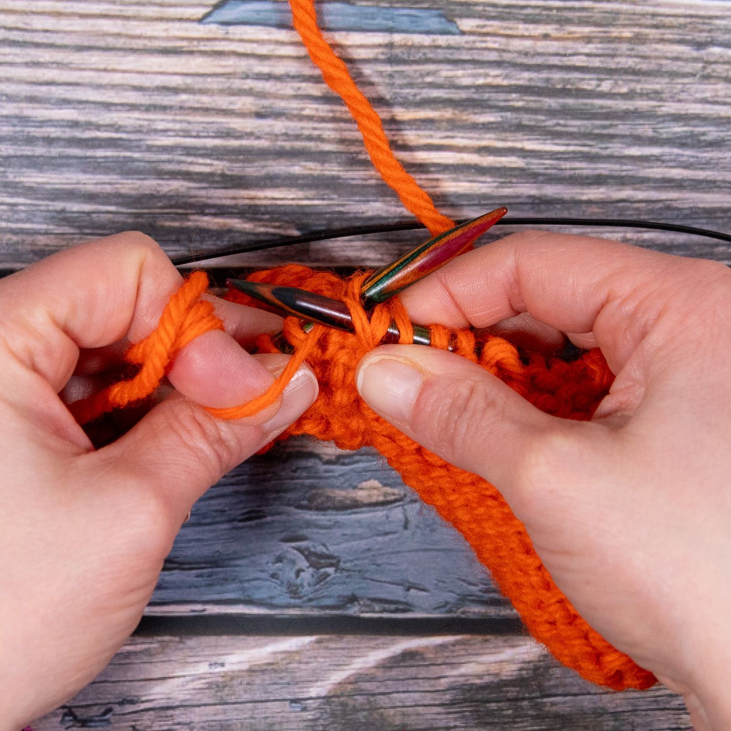 Hände stricken mit orangefarbenem Garn und Stricknadeln auf einer Holzfläche.