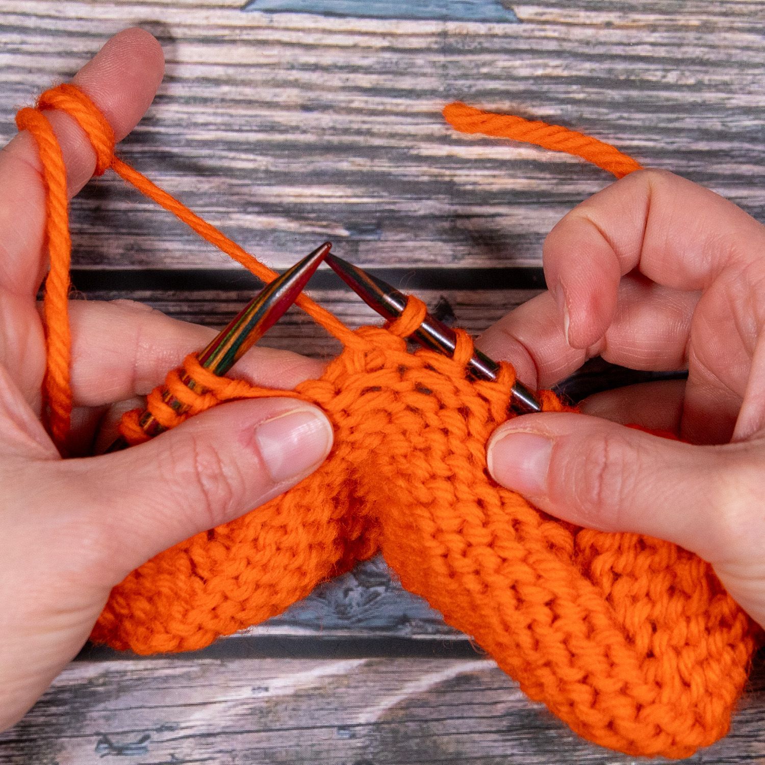 Hände stricken mit orangefarbenem Garn; Nadeln, Maschen und fertiges Strickstück sind sichtbar.