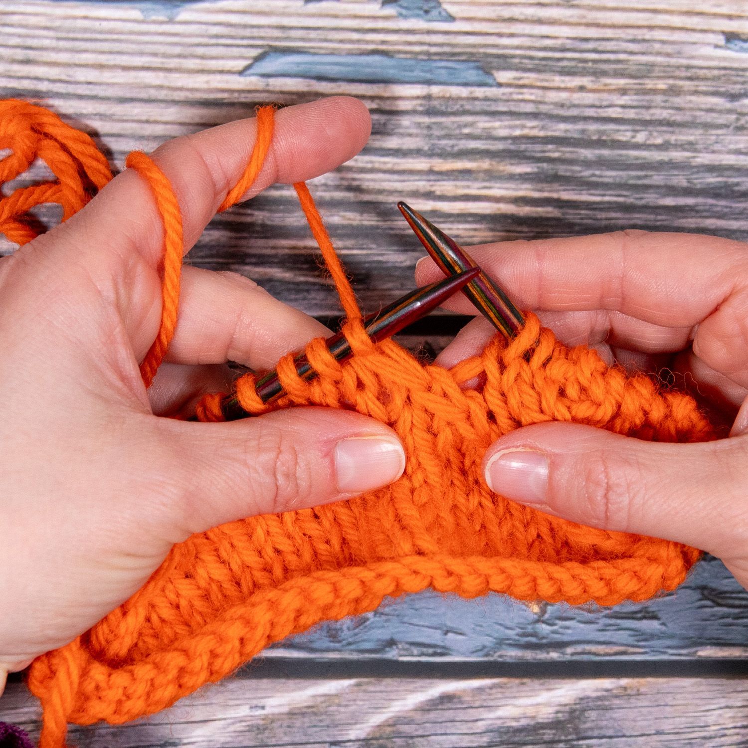 Hände stricken mit orangefarbenem Garn auf einer Holzoberfläche.