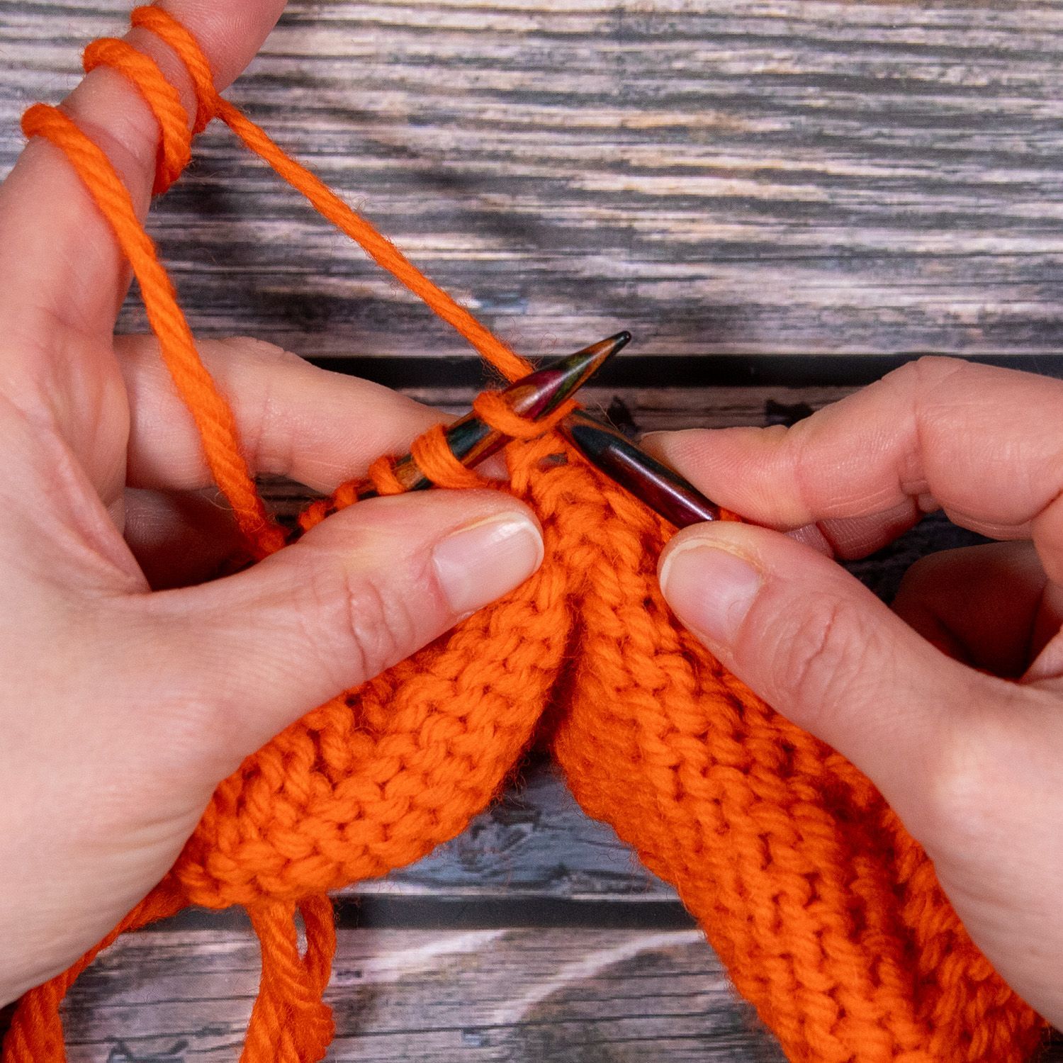 Hände stricken mit orangefarbenem Garn; Nadeln werden in die Maschen eingeführt.