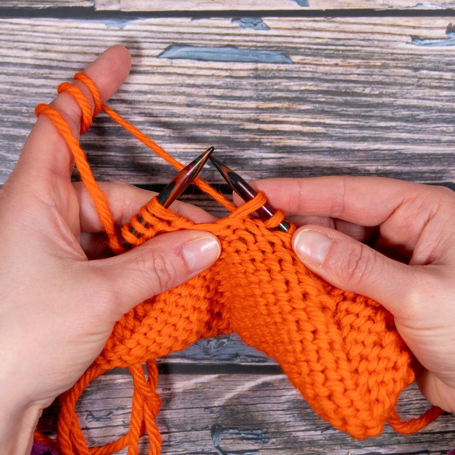 Hände stricken mit orangefarbenem Garn; Nadeln stecken im Stoff.