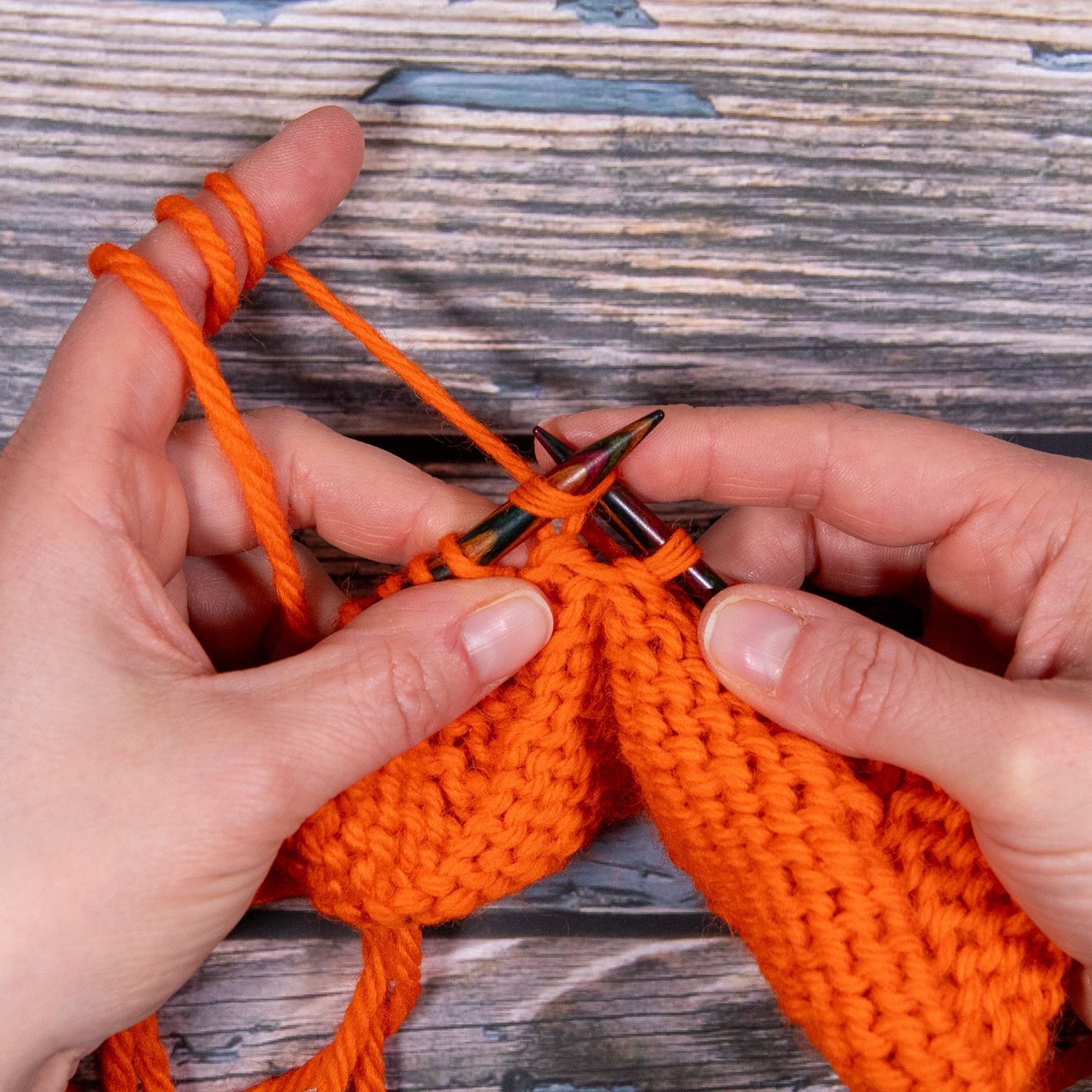 Hände stricken mit orangefarbenem Garn; Nadeln, eine teilweise fertiggestellte Reihe rechter Maschen.