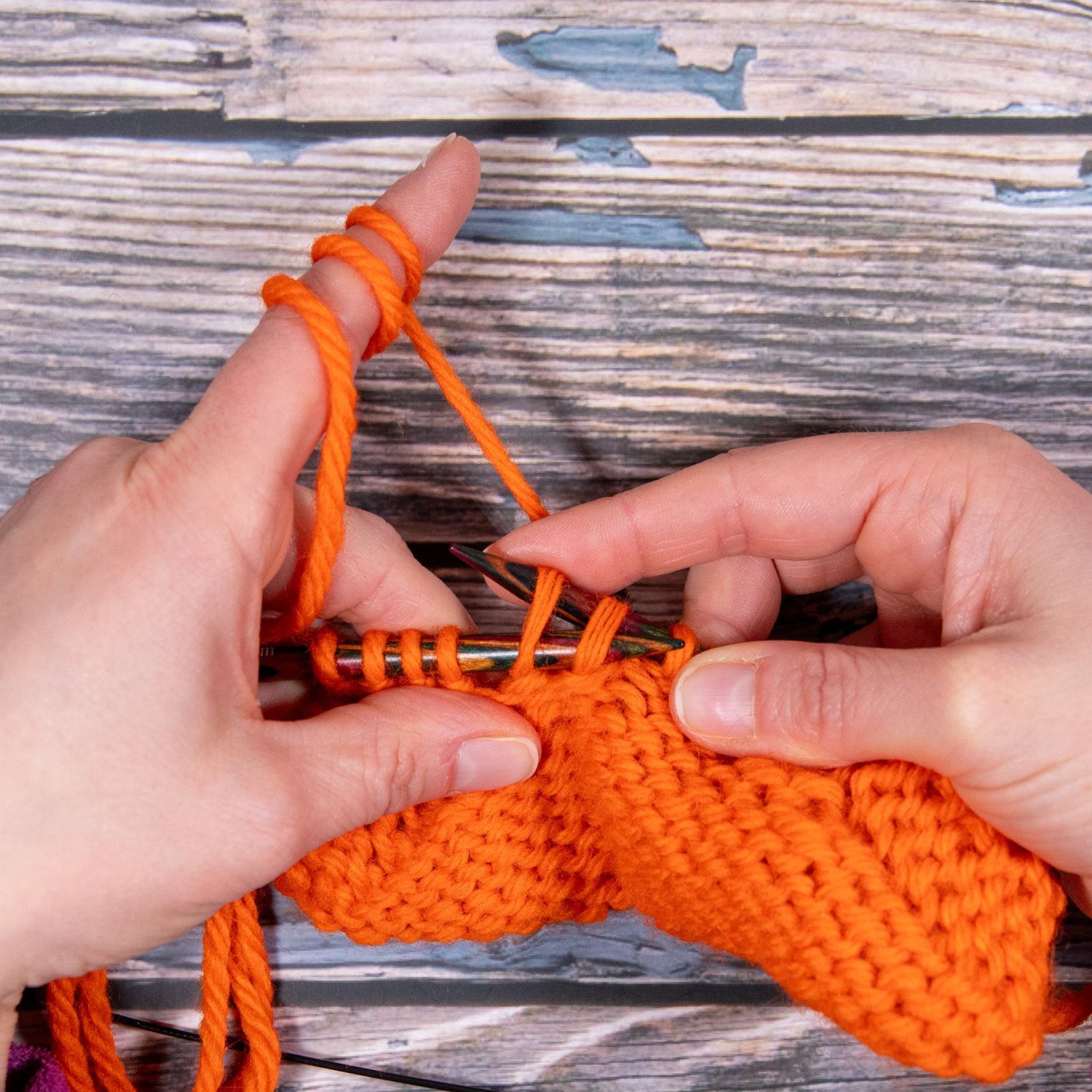 Hände stricken mit orangefarbenem Garn und zeigen den Vorgang, eine Schlaufe durch eine Masche zu ziehen.