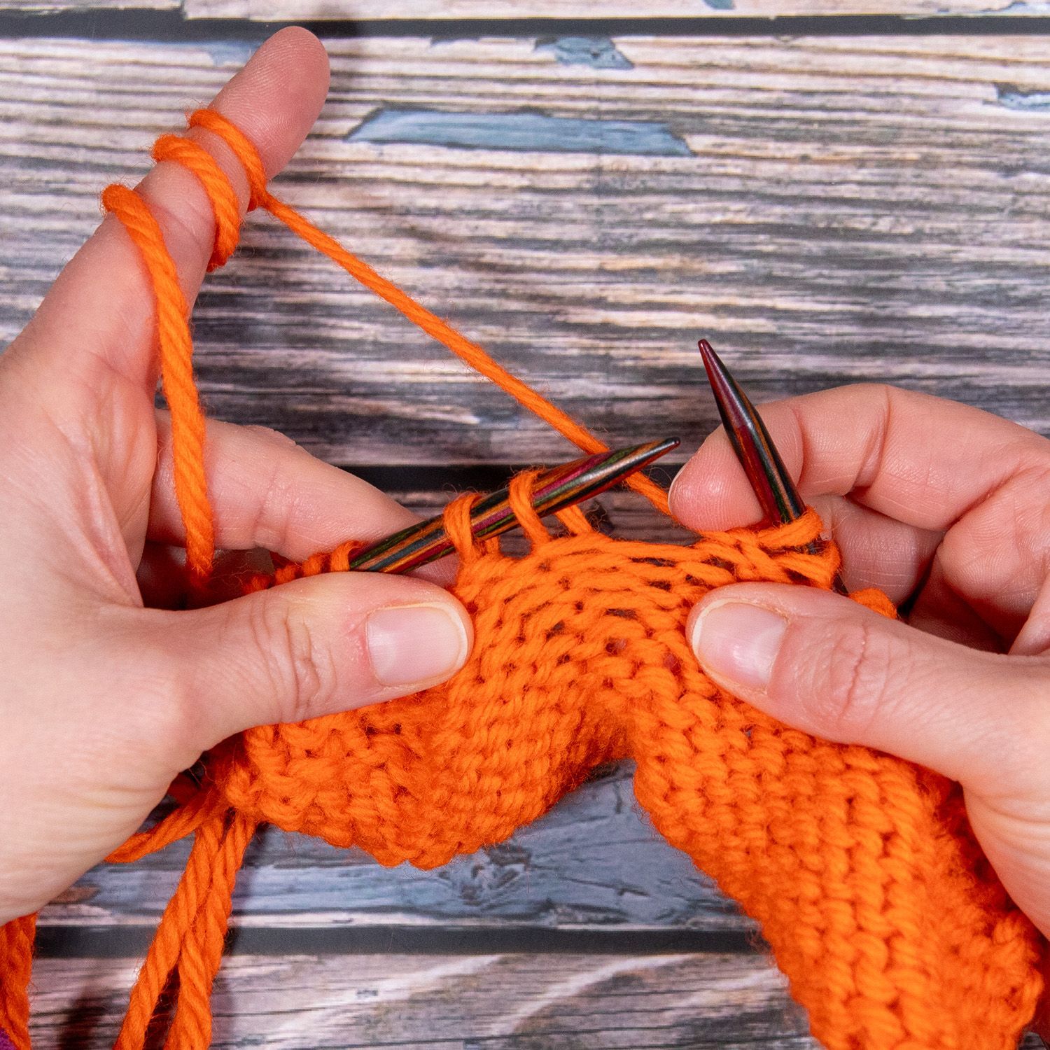 Hände stricken mit orangefarbenem Garn und zeigen den Vorgang des Maschenbildens auf den Nadeln.