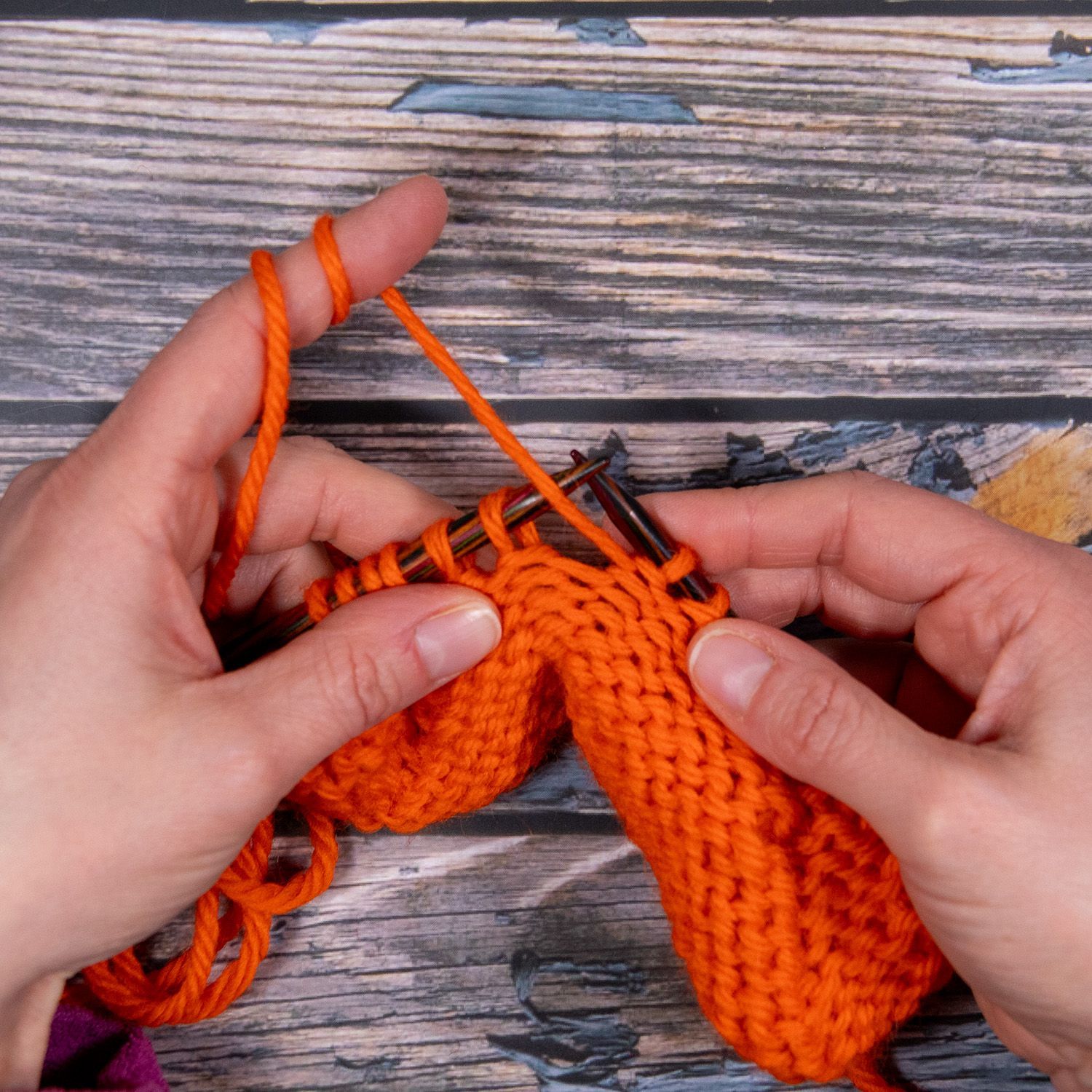 Hände stricken mit orangefarbenem Garn auf einer Holzfläche.