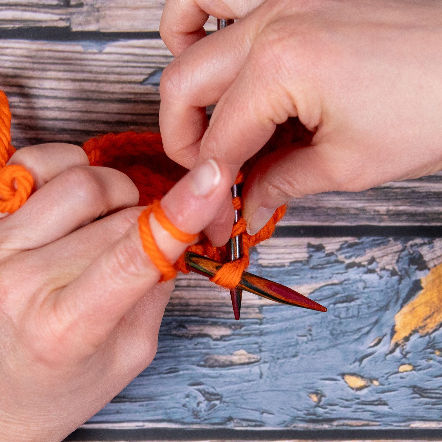 Hände stricken mit orangefarbenem Garn und Holznadeln auf einem blaugrauen Untergrund.