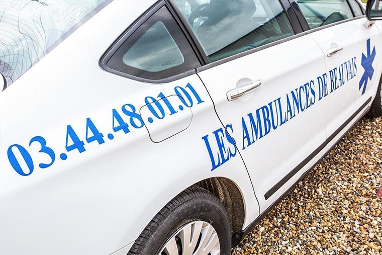 Ambulance blanche avec inscription bleue et emblème en forme d'étoile, stationnée à l'extérieur. Numéro de téléphone : 03.44.48.01.01.