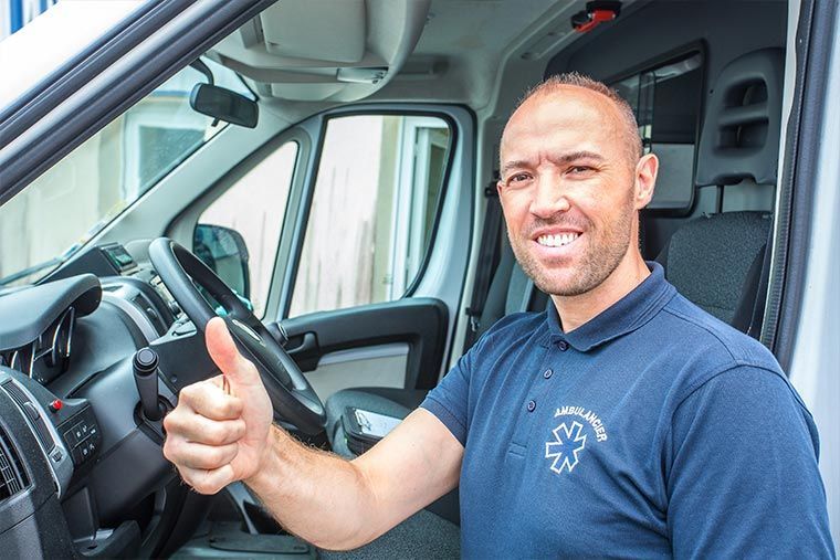 Un homme dans une ambulance, pouce levé. Il porte une chemise bleue avec le logo des services d'urgence et sourit.