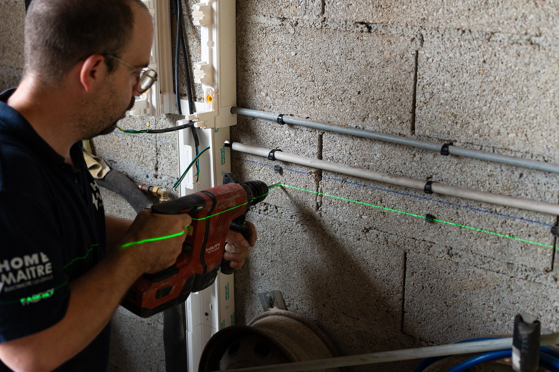 Un homme perce un mur en béton à l'aide d'un niveau laser, des tuyaux étant fixés à proximité.