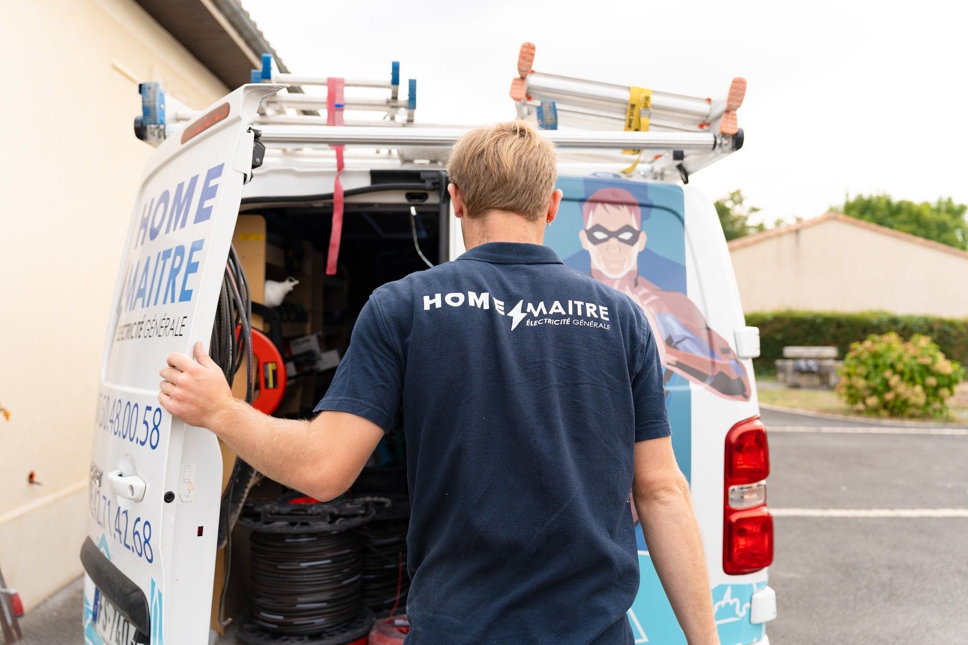 Un homme en chemise bleu marine ornée du logo d'une entreprise ouvre l'arrière d'une camionnette de service ; des outils sont visibles à l'intérieur.