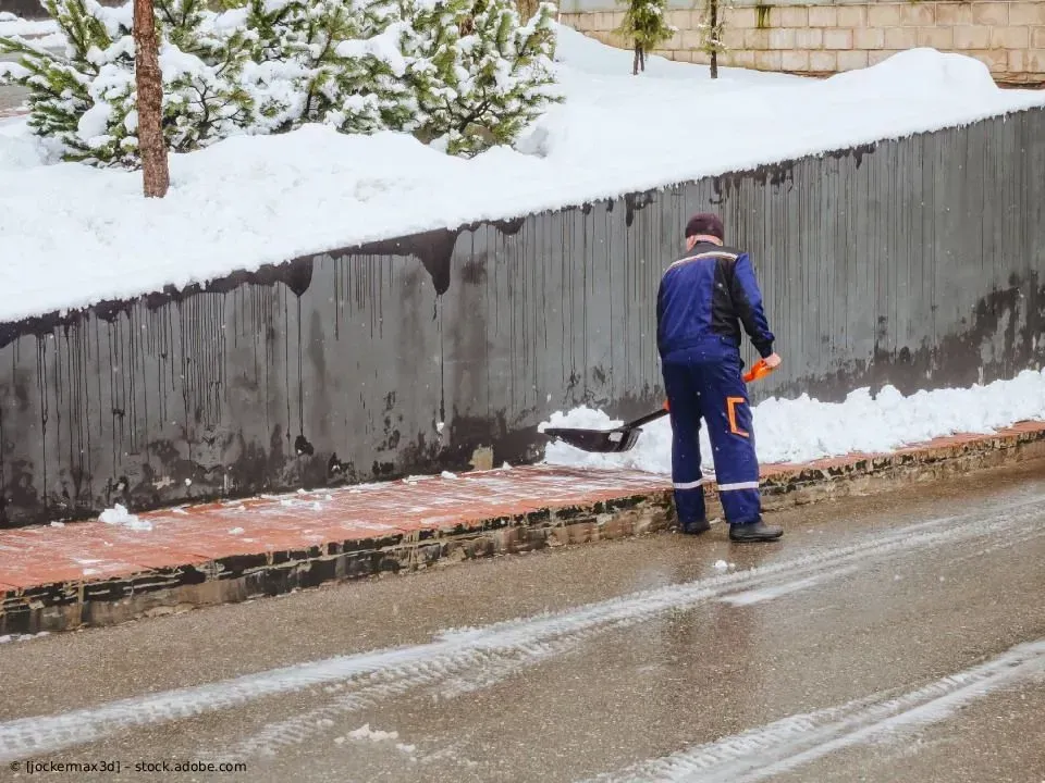 Ein Arbeiter in blauer Winterkleidung schaufelt Schnee neben einer Straße und einer Stützmauer.