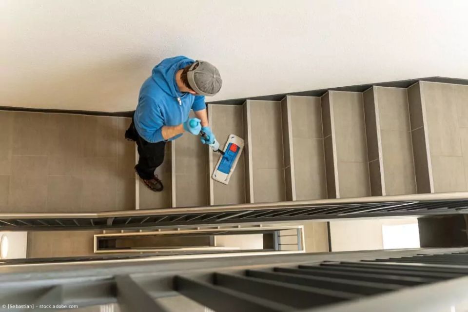 Ein Arbeiter in blauer Jacke reinigt von oben ein Treppenhaus mit einem Mopp und einem Eimer.