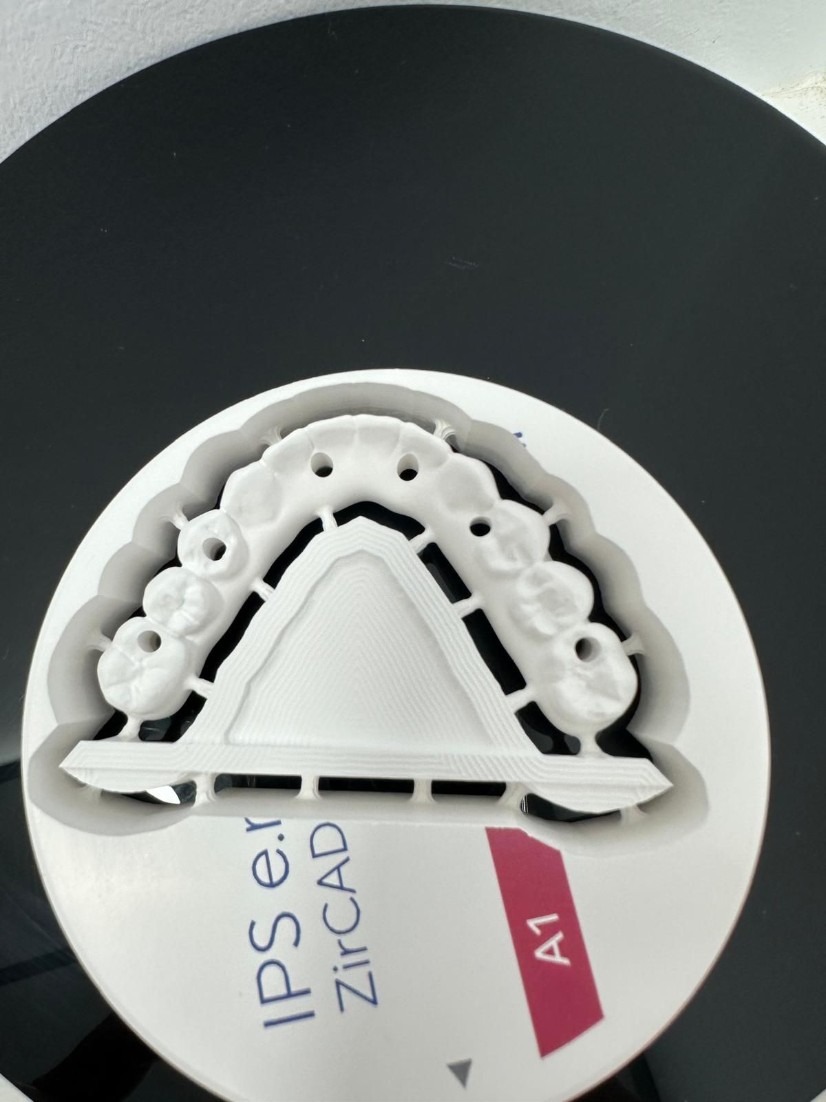 Un puente dental de zirconio blanco fresado (tono A1) sobre su disco de fabricación original.