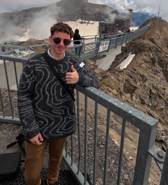 Una persona con gafas de sol hace un gesto de aprobación con el pulgar en un mirador de montaña, junto a una barandilla, con vapor y terreno rocoso al fondo.