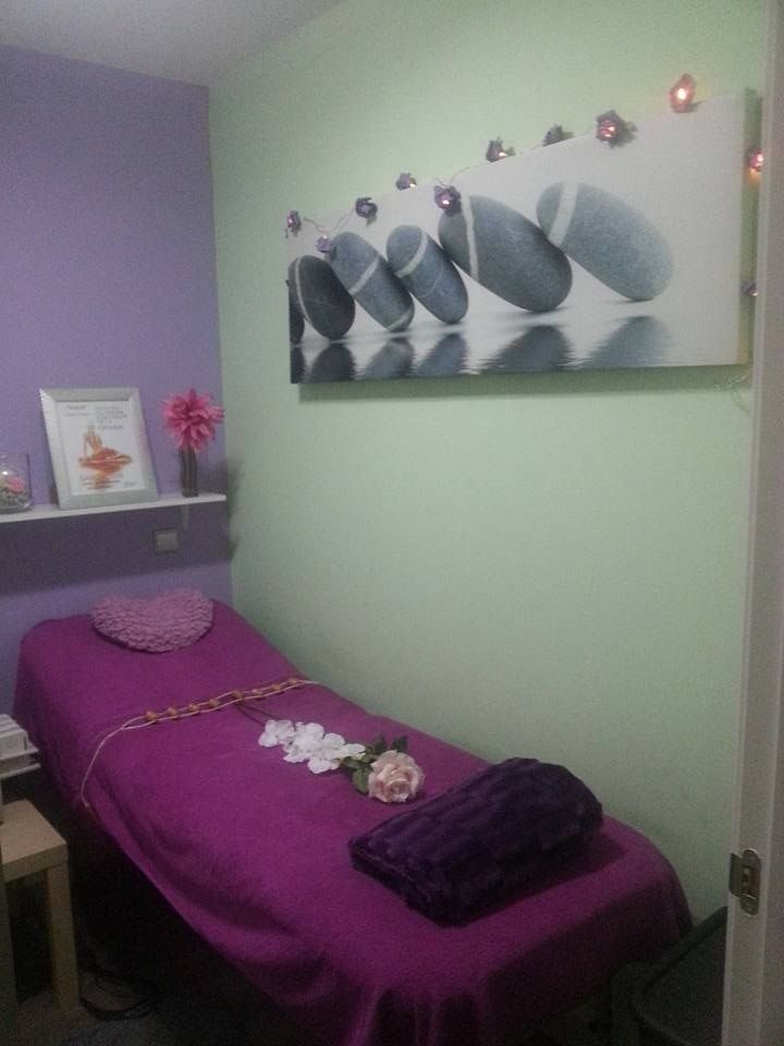 Pequeña sala de masajes con camilla de tratamiento morada, pared color lavanda, estante y arte abstracto en la pared.