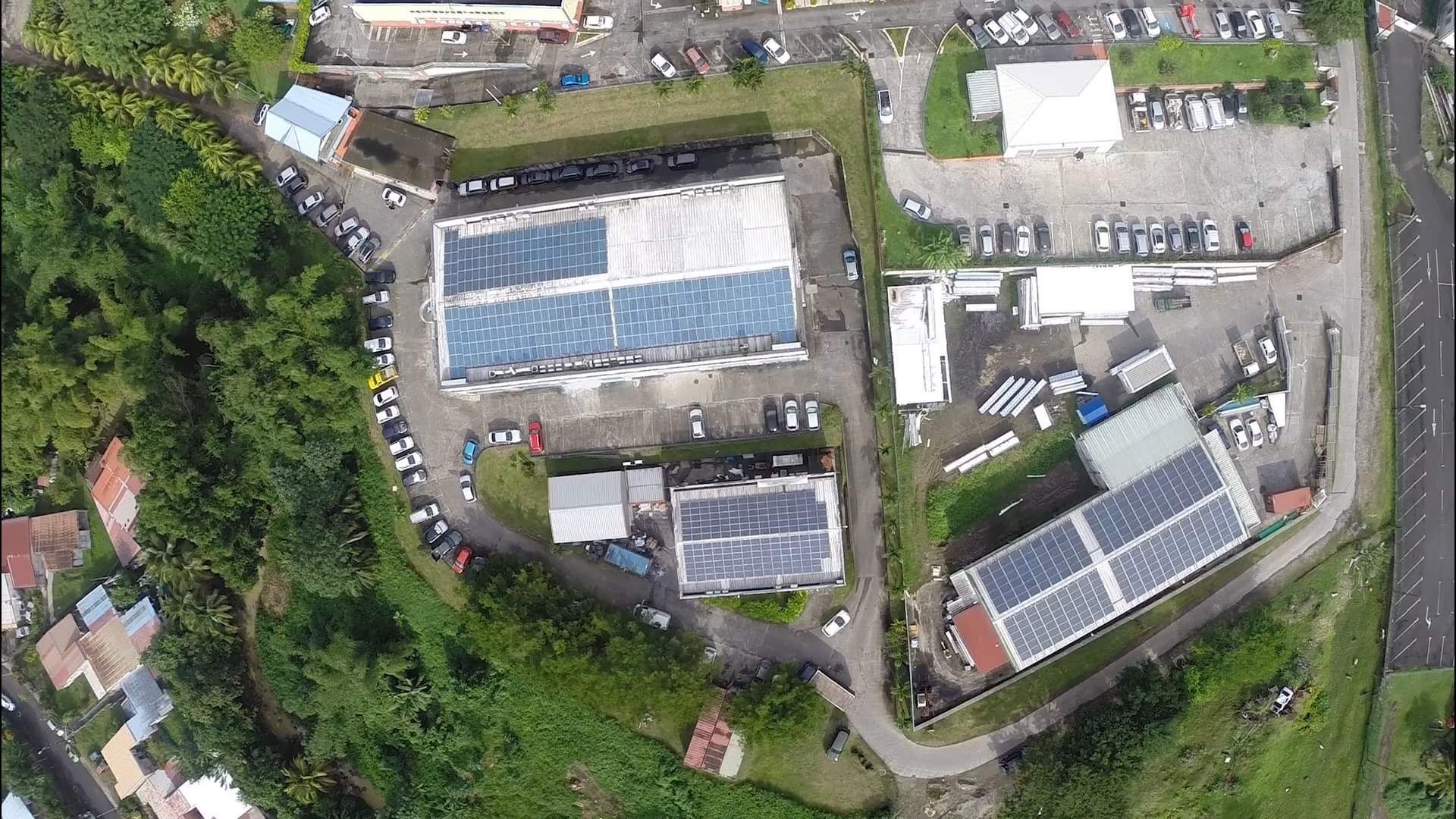 Vue aérienne : bâtiments avec des panneaux solaires sur les toits, entourés de voitures garées et d'une végétation luxuriante.