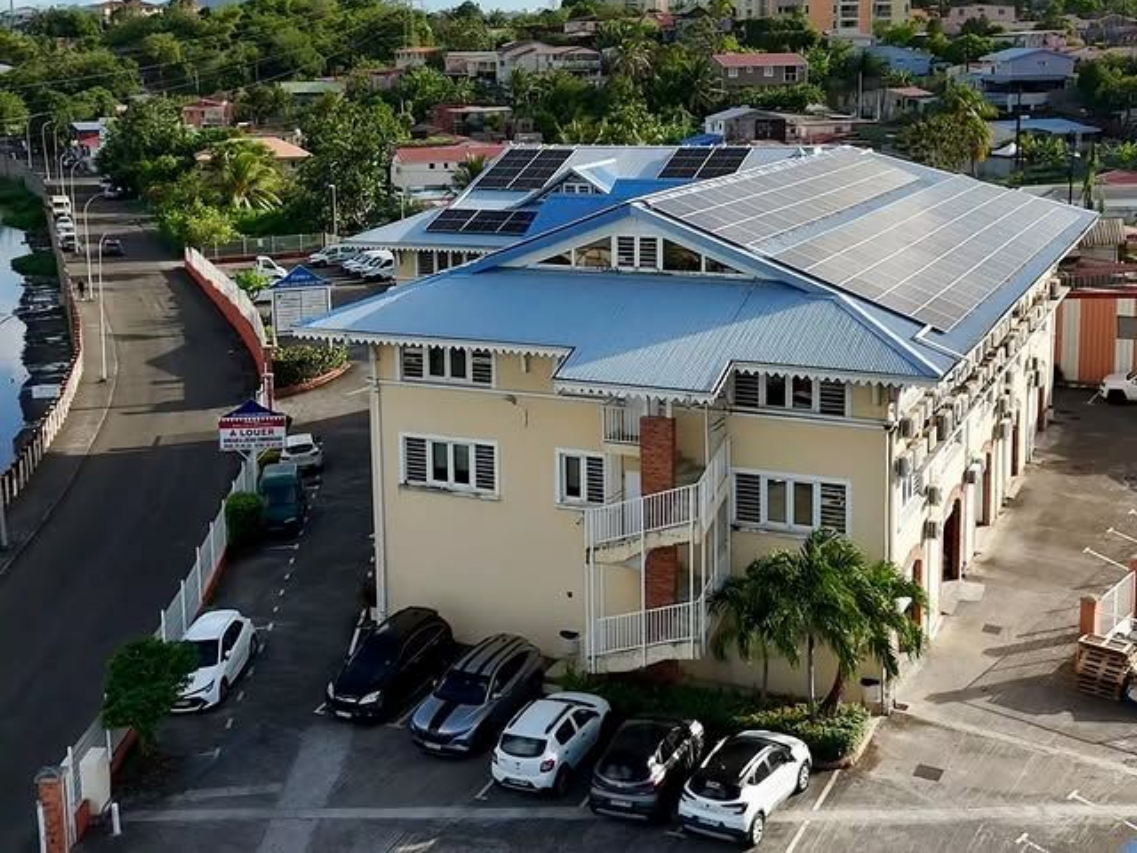 Bâtiment beige au toit bleu, panneaux solaires et voitures garées, au bord d'un canal, dans un cadre ensoleillé.
