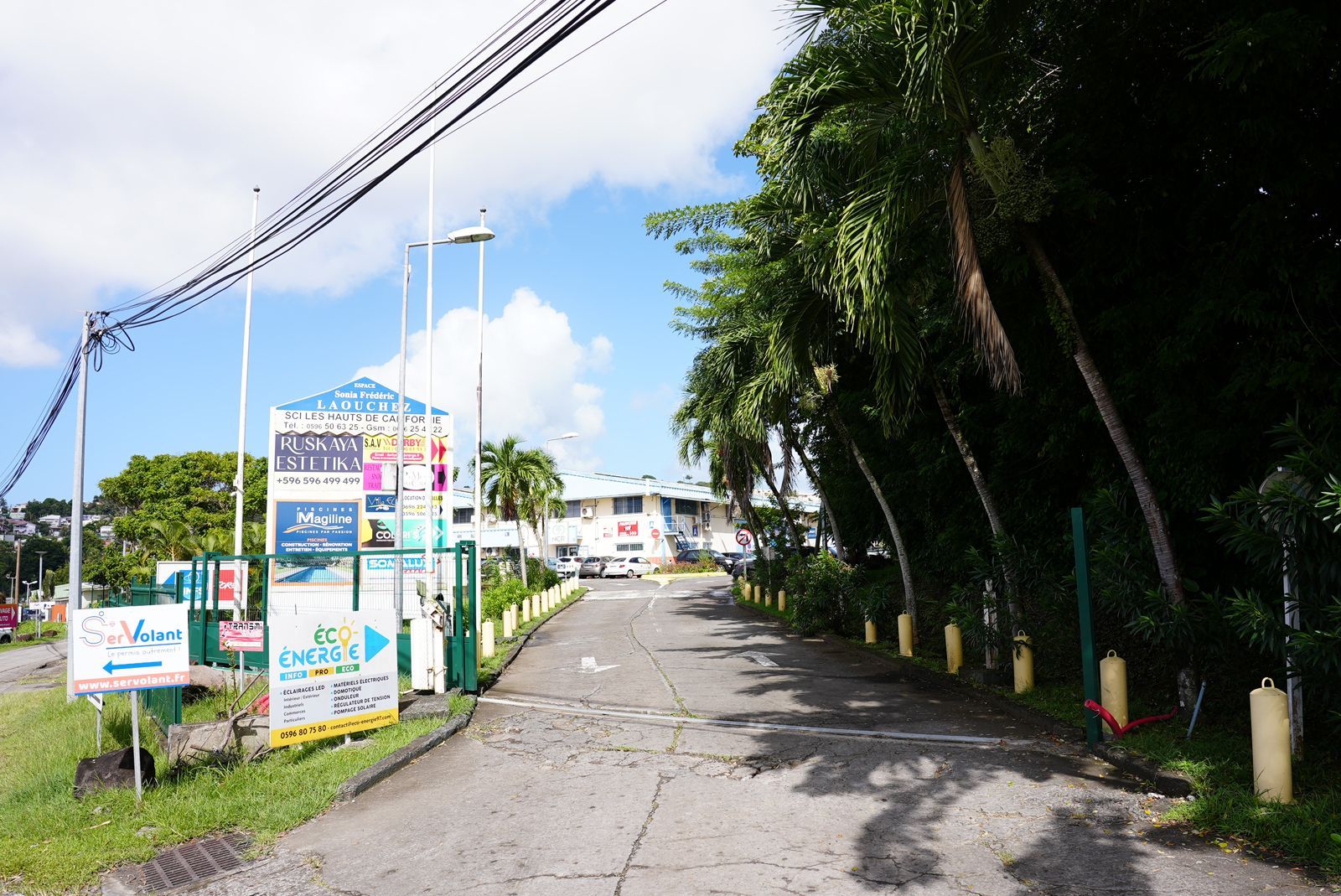 Entrée d'une zone commerciale avec panneaux, portail et palmiers sous un ciel bleu vif.