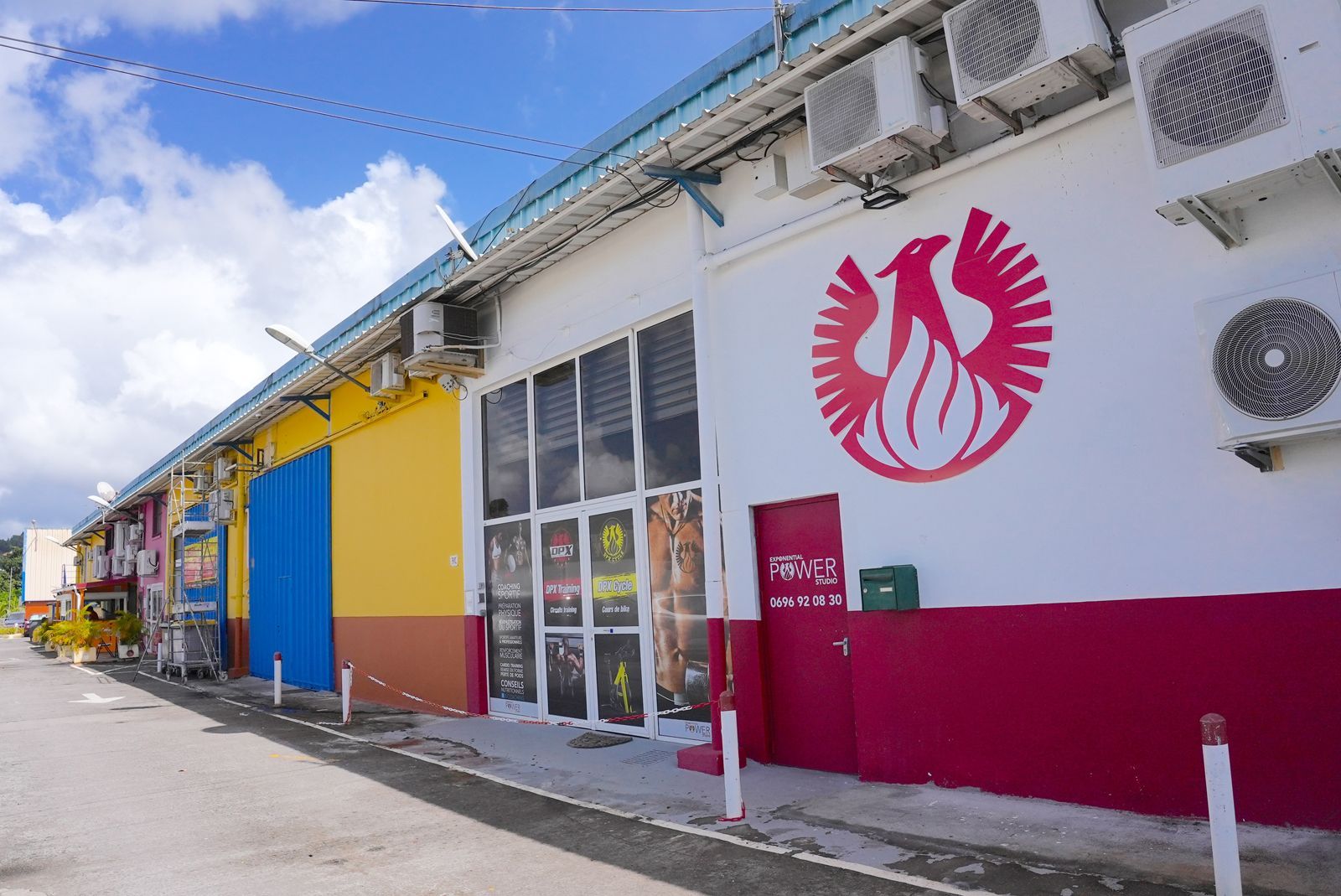 Rangée de devantures de magasins peintes de couleurs vives avec un grand logo de phénix rouge sur un mur blanc, sous un ciel bleu.