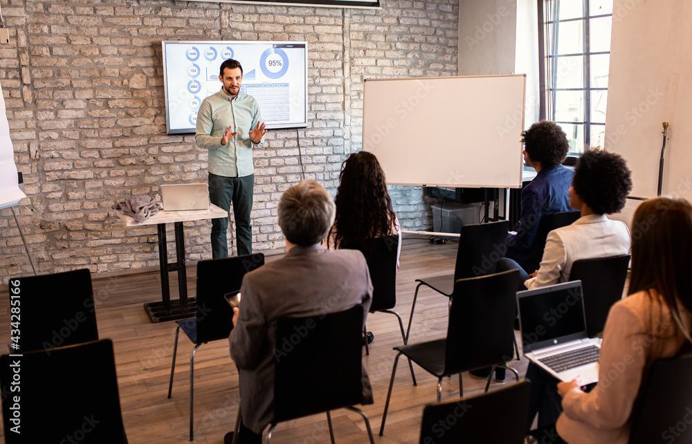 Un homme présente des données à un public dans une salle aux murs de briques. Les participants sont assis, certains équipés d'ordinateurs portables.