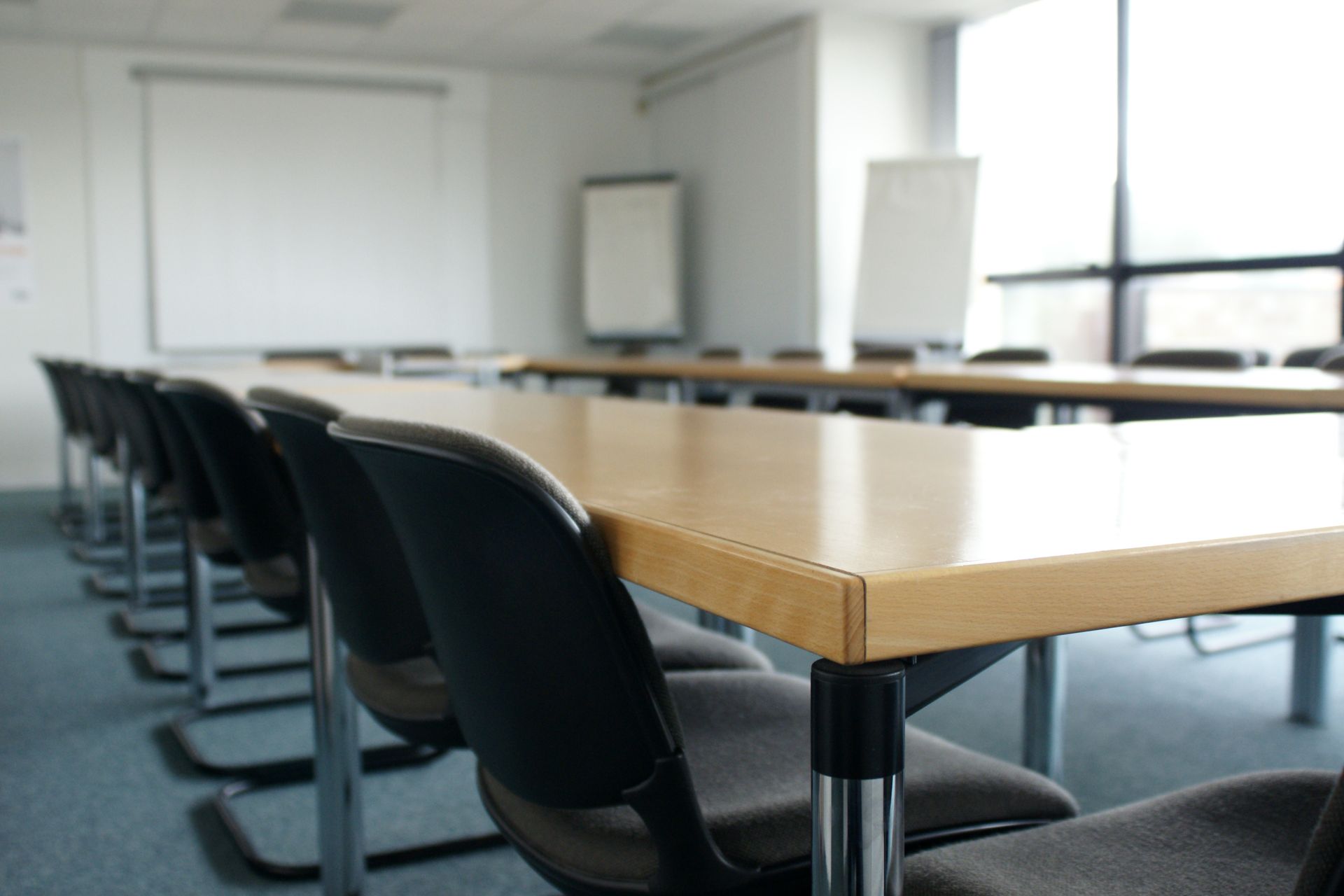 Salle de conférence vide avec de longues tables, des chaises noires et des tableaux blancs.