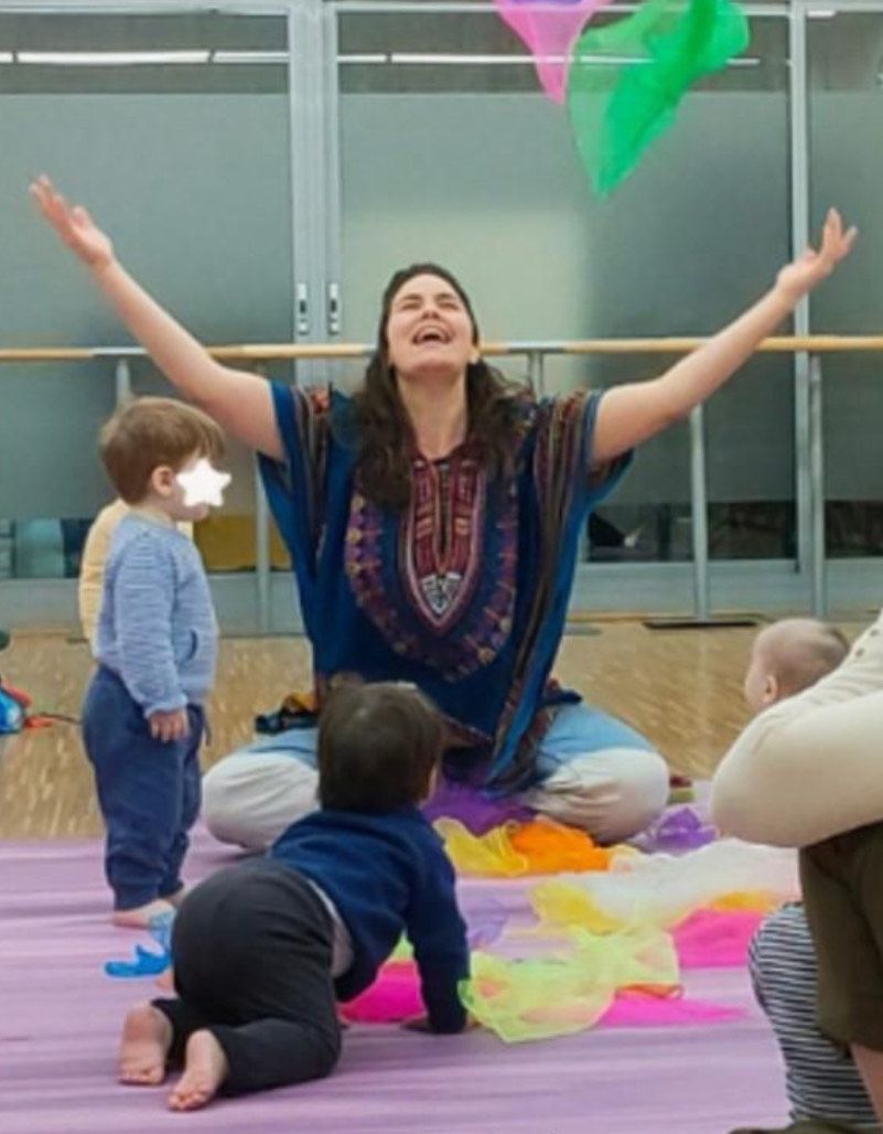 Mujer con los brazos en alto, liderando una actividad infantil con bufandas de colores y niños en un espacio de juegos.