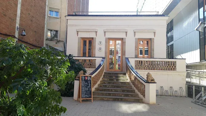Un edificio blanco con unos escalones que conducen a unas puertas de madera. Un cartel a la izquierda con vegetación en primer plano.