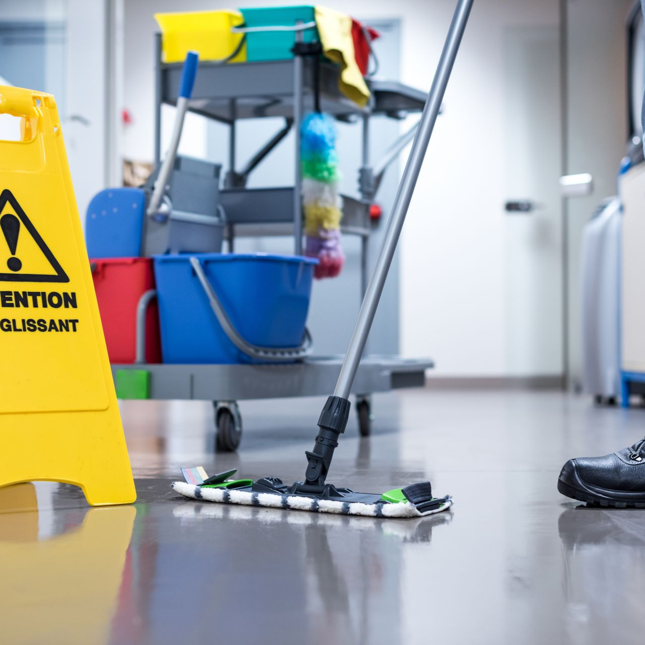Une personne lave un sol mouillé à côté d'un panneau de signalisation jaune et d'un chariot de nettoyage dans un couloir intérieur.