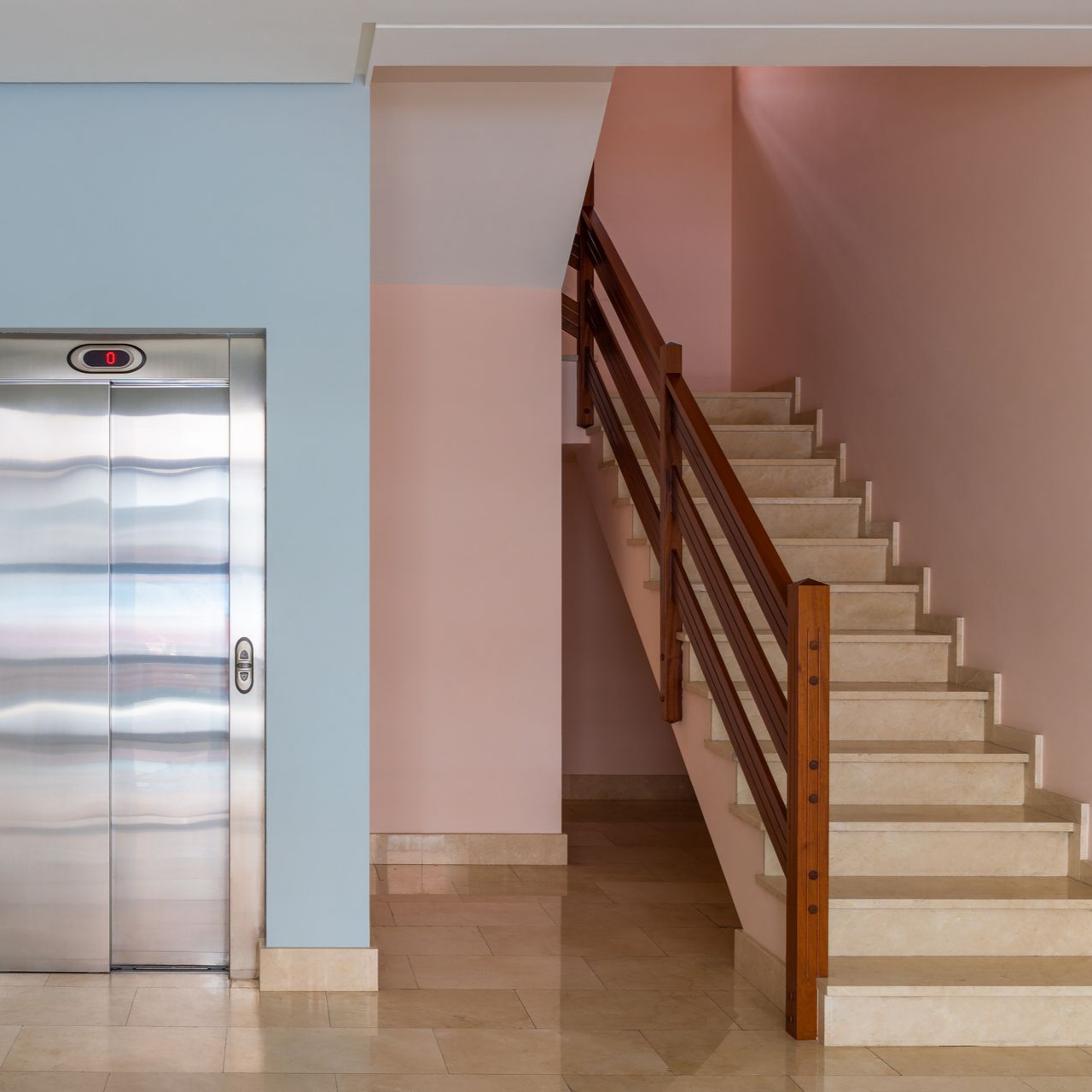 Escalier à côté d'un ascenseur dans le hall d'un immeuble aux sols carrelés beiges et aux murs bleu et rose pastel.