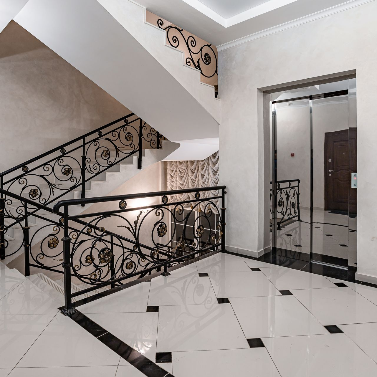 Escalier intérieur avec rampes noires ouvragées et sol carrelé blanc brillant, à côté d'un ascenseur.