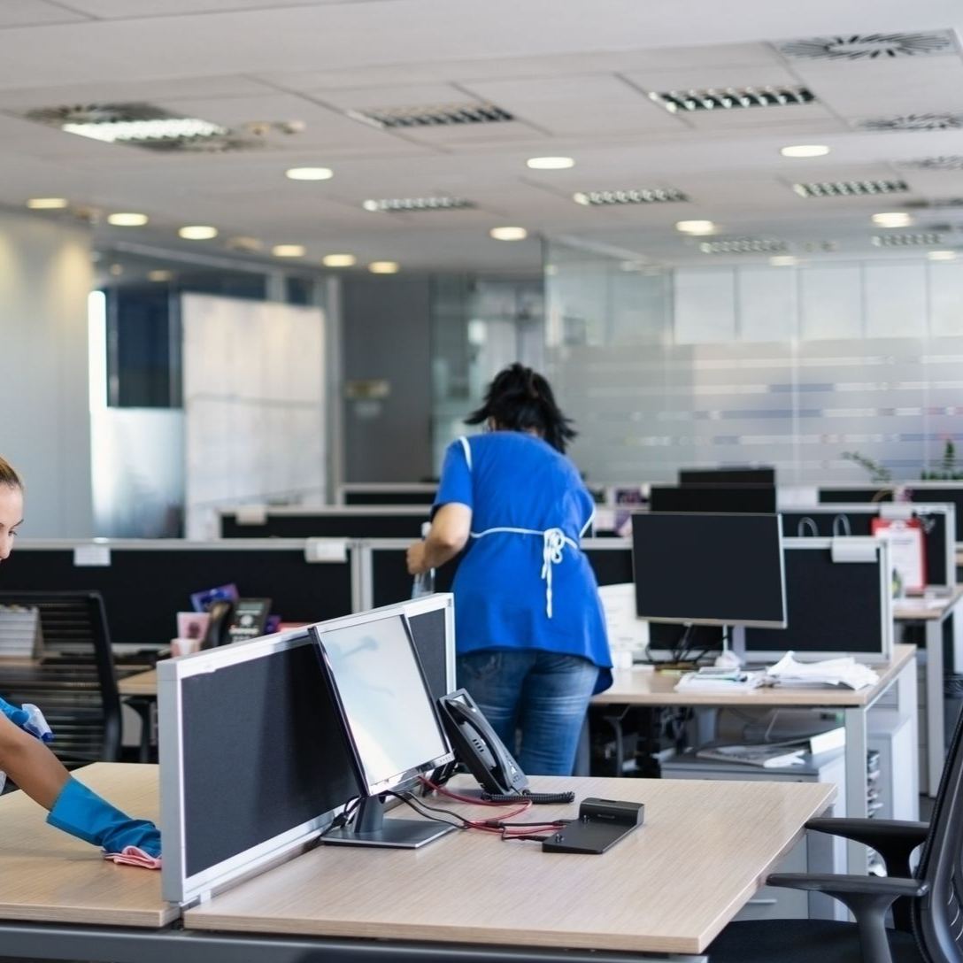 Deux personnes en uniforme bleu et gants nettoient des bureaux dans un espace de travail ouvert et lumineux.