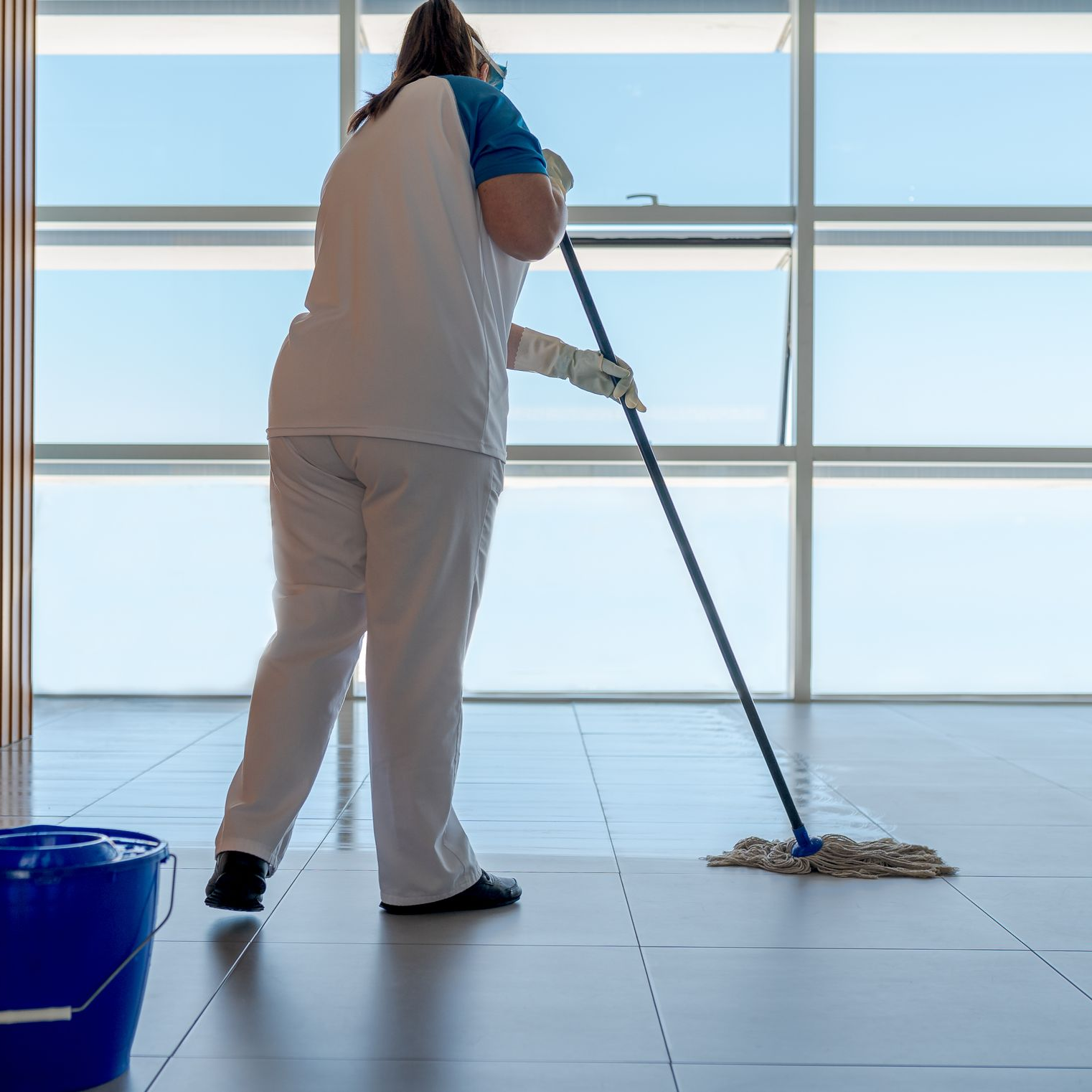 Une personne en uniforme blanc et bleu lave un sol carrelé et lumineux devant de grandes fenêtres, un seau bleu se trouvant à proximité.