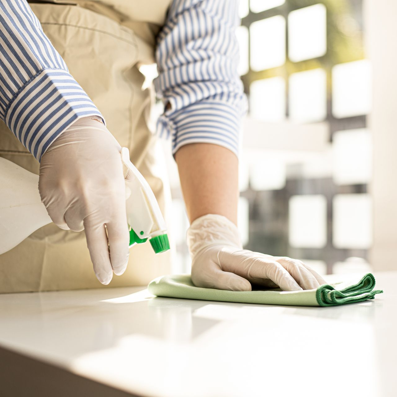 Une personne portant des gants blancs et une chemise à rayures nettoie une surface blanche avec un vaporisateur et un chiffon vert.