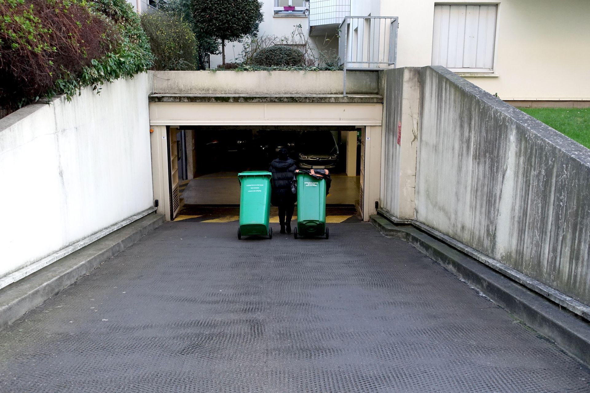 Une personne descend une allée en pente menant à un parking souterrain en tirant deux poubelles vertes à roulettes.