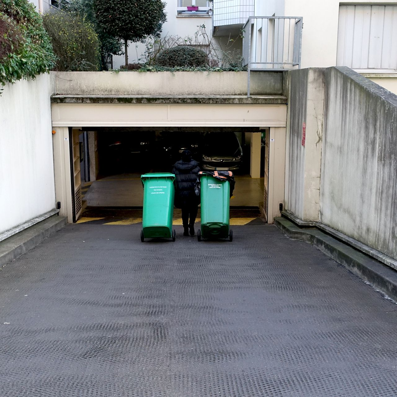 Une personne vêtue de sombre fait rouler deux poubelles vertes sur une allée en béton jusqu'à un parking souterrain.